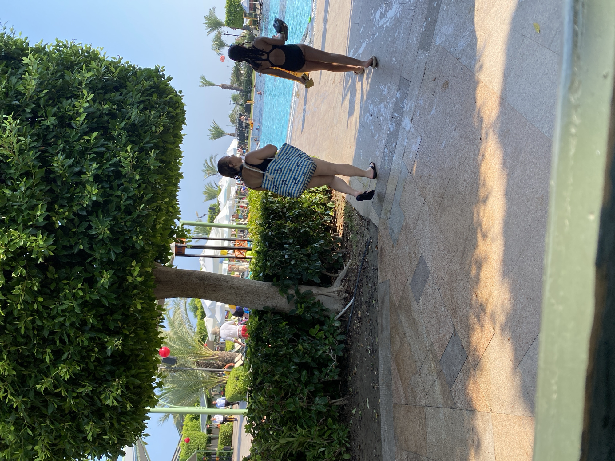 Two females, one adult and one child, walk away from the camera on a tiled pathway next to a swimming pool. The adult, on the left, wears a black swimsuit and carries a large blue and white striped tote bag. The child, on the right, wears a black swimsuit and carries a dark object in her right hand. Palm trees and white umbrellas are visible around the pool. The location is Aksu, Turkey.