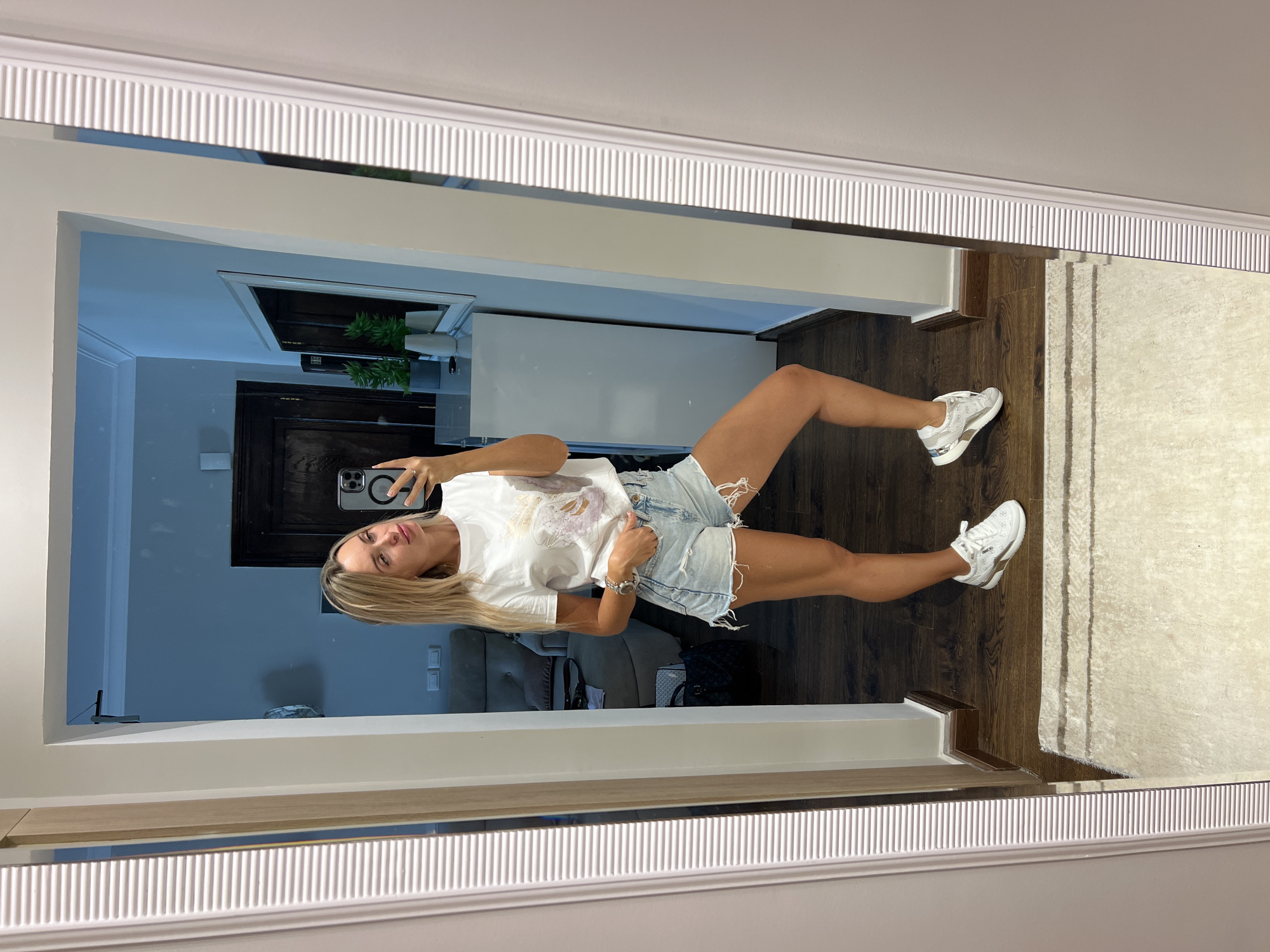 A blonde woman in her late 20s or early 30s is taking a mirror selfie in a hallway. She is wearing a white t-shirt with a graphic print, light blue denim shorts, and white athletic shoes. She has one hand in her pocket and is holding her phone in the other. The hallway has a wooden floor, a large mirror with a ribbed frame, and a white carpet in the foreground. In the background, visible in the mirror, is a room with a grey sofa, a dark wooden door, and a white dresser with plants on top. The lighting suggests it is daytime or well-lit interior.