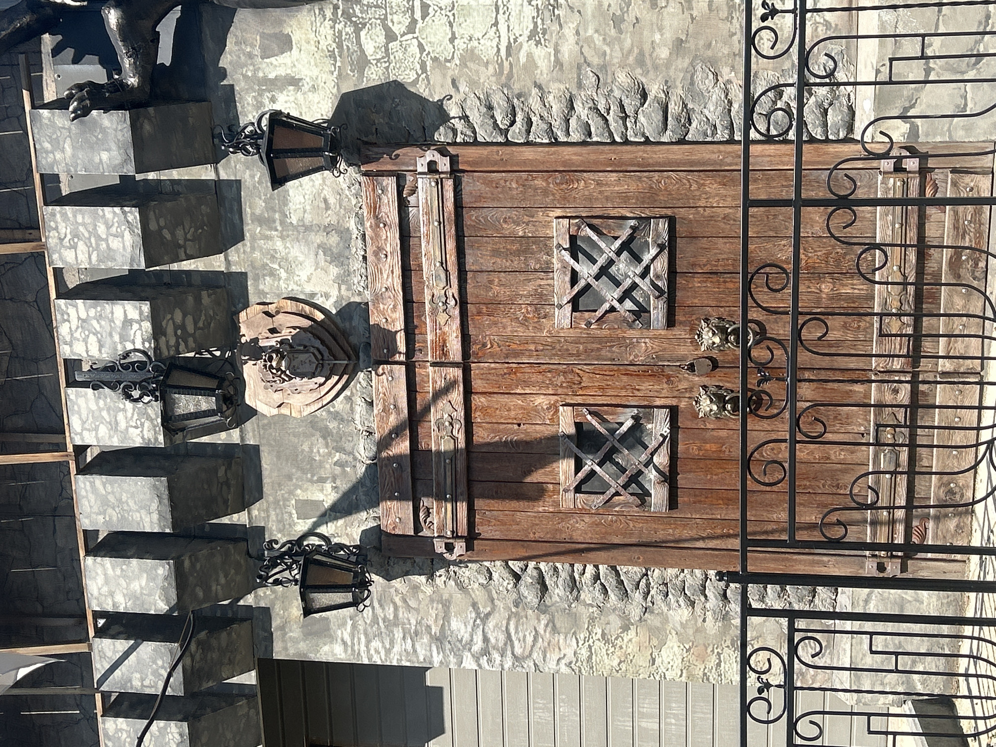 The image shows the entrance to a building with a medieval, castle-like aesthetic. The main feature is a large, rustic wooden double door with two small, square windows, each fitted with a criss-cross metal grate. Above the door, mounted on the stone facade, is a shield-shaped emblem with a crest. Two ornate, dark metal lanterns hang from the wall on either side of the emblem, and a similar lantern is visible to the left of the door. The building's exterior is constructed of rough stone and features decorative battlements along the top. A black wrought-iron fence with decorative scrollwork is positioned in front of the door. The overall impression is one of a themed attraction or establishment, potentially a restaurant, bar, or entertainment venue.
This building is located at 115 Komsomolsky Prospekt in Perm, Russia. It is known as the "Razgulyai" complex, a popular entertainment venue with a medieval theme.
