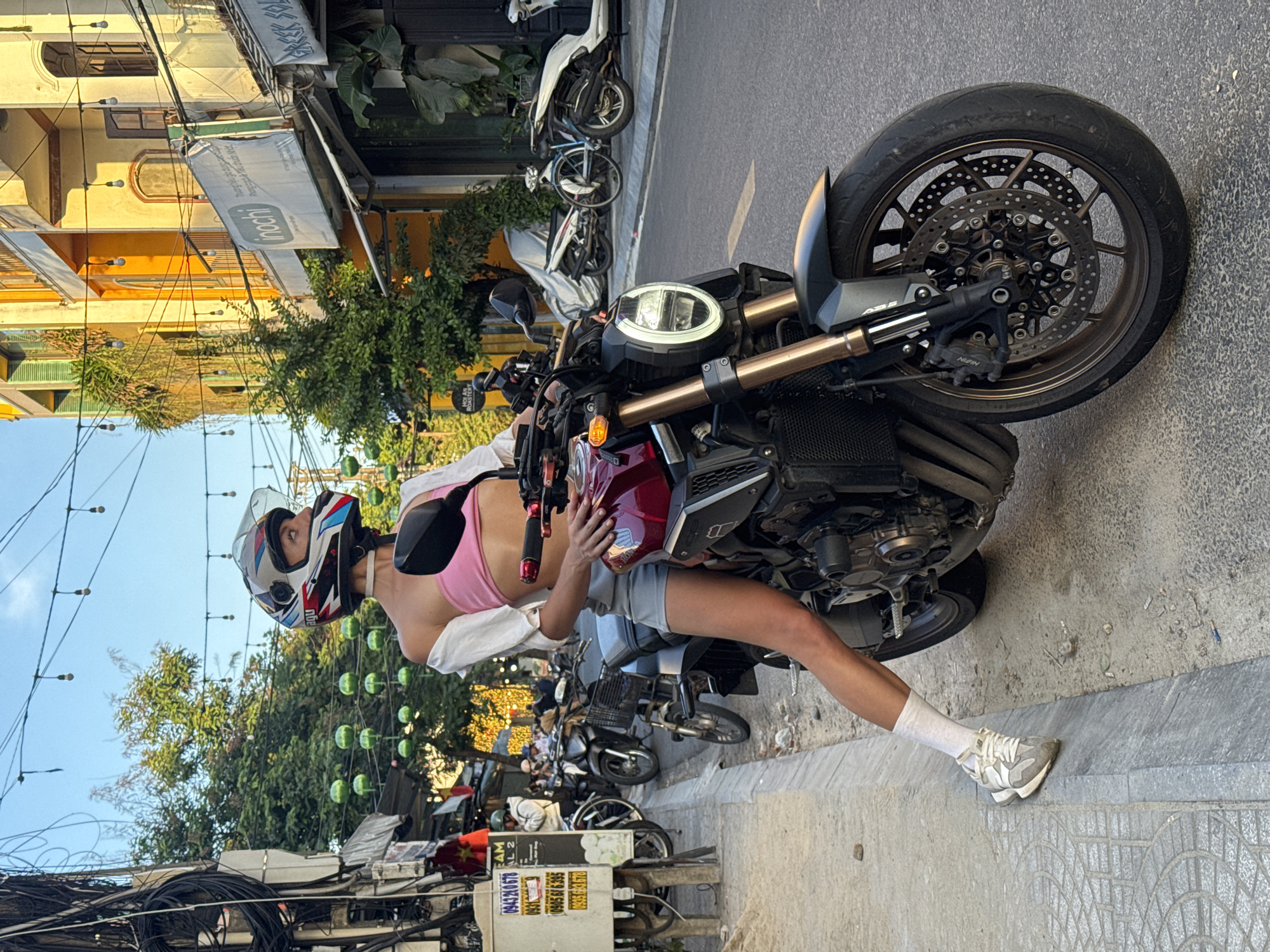 A person wearing a helmet, pink top, shorts, white shirt, white socks, and white sneakers is perched on a burgundy and black motorcycle on a street in Hội An Ward, Vietnam. The motorcycle is angled slightly towards the viewer with its front tire close to the edge of the sidewalk. The background features buildings, trees, motorbikes, and an array of cables and wires overhead. A sign with Vietnamese writing can be seen near the bottom of the image. The lighting is bright, suggesting daytime.