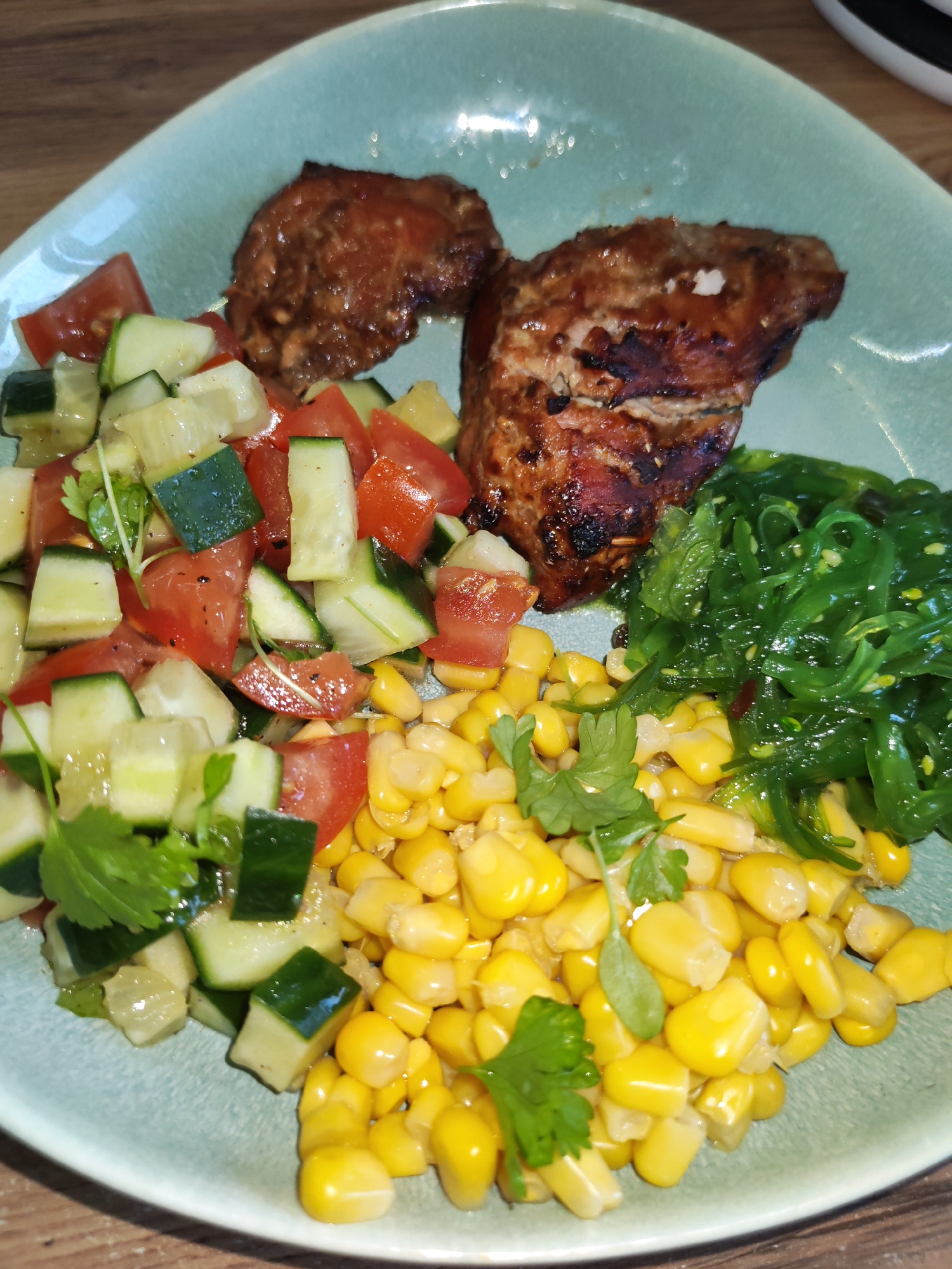 This close-up shot displays a meal served on a light blue-green ceramic plate, resting on a wooden table. The plate is filled with a variety of colorful dishes, suggesting a balanced and appealing meal.
The main subject is the food itself, arranged in distinct sections on the plate. To the upper right, there are two pieces of grilled meat, possibly pork or chicken, with visible char marks and a glossy, dark marinade, indicating they are well-cooked.
On the left side of the plate is a fresh salad composed of diced cucumbers and tomatoes, interspersed with green sprigs of what appears to be cilantro or parsley, and a light dressing. The cucumbers show their green skin and paler flesh, while the tomatoes are a vibrant red.
Below the grilled meat and to the right of the cucumber and tomato salad, there's a pile of bright yellow sweet corn kernels, some also garnished with a sprig of cilantro or parsley. Adjacent to the corn and next to the grilled meat is a serving of glossy, vibrant green seaweed salad, likely wakame, which appears lightly dressed and possibly contains sesame seeds.
The meal is presented neatly, with each component distinct yet harmoniously arranged. The overall impression is that of a freshly prepared, healthy, and flavorful dish. There are no people visible in the image, nor any discernible text. The setting is indoors, indicated by the wooden table surface, likely a dining area or kitchen. The time of day cannot be determined, and there are no specific location cues visible within the image that point to Izhevsk, Russia, beyond the general context provided.
