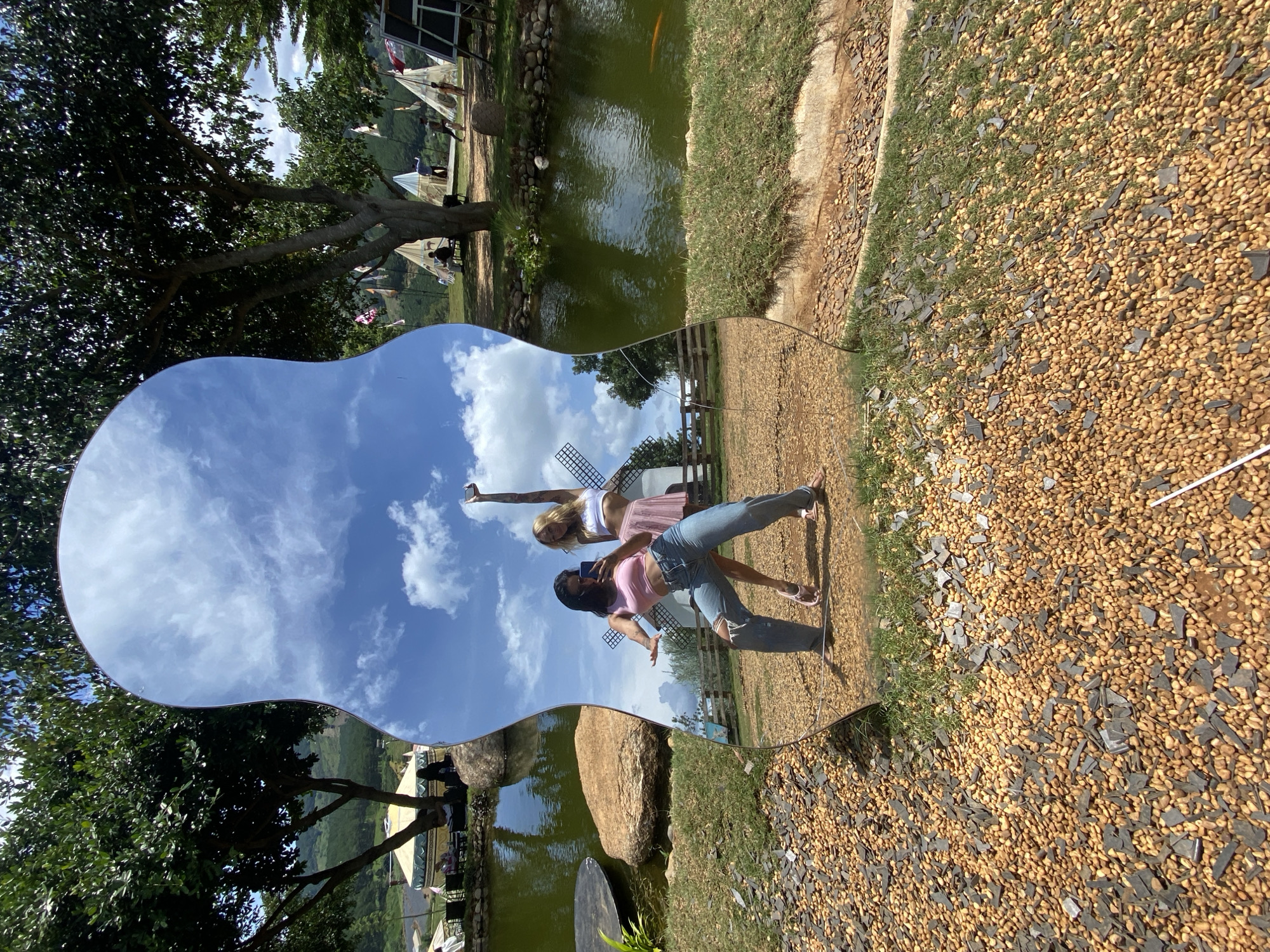 This media file captures a playful moment at a scenic outdoor location in Thôn 5, Vietnam. The primary subjects are two young women posing for a photo. One woman, with blonde hair, is captured in mid-air, her arms outstretched and holding a smartphone to take a selfie. She is wearing a white crop top and a pink skirt. The other woman, with dark hair, is leaning forward, looking at the phone, and is dressed in a pink crop top and distressed blue jeans.
The setting appears to be a park or resort, characterized by a pond with green water, large rocks, and a path covered in gravel and grass. In the reflection, a Dutch-style windmill can be seen, adding a unique element to the landscape. Trees with lush green foliage frame the scene, and in the distance, rolling hills suggest a rural or mountainous environment. The sky is a vibrant blue with fluffy white clouds, indicating fair weather and a bright, sunny day, likely late morning or early afternoon. The overall mood is cheerful and energetic, with the women appearing to be enjoying their time together. The reflection in the mirror distorts the environment, creating an interesting visual effect that overlays the real-world scene with its mirrored counterpart. There is no visible text in the media file.