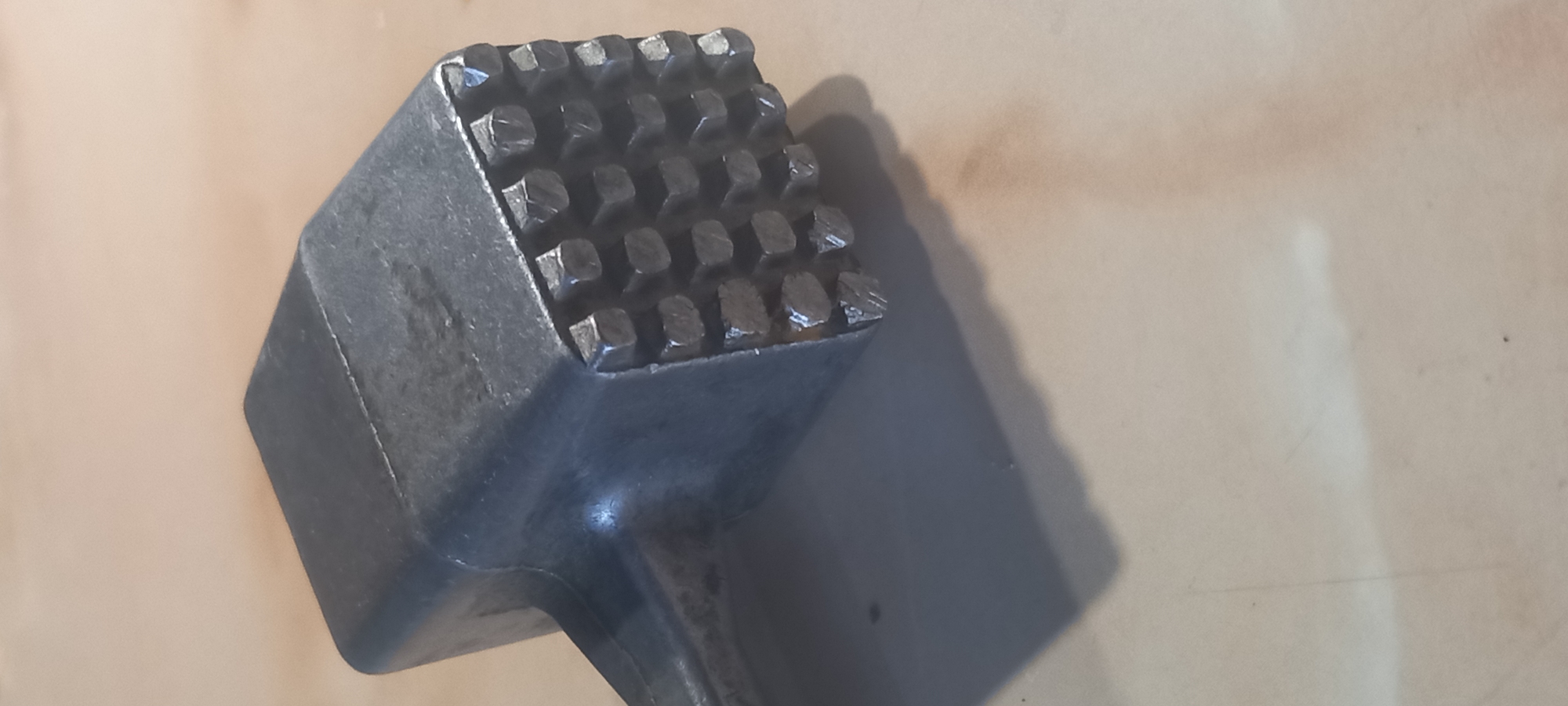 The image shows a meat tenderizer lying on a light-colored surface. The tenderizer is made of a dark metal, possibly cast iron or aluminum, and has a grid of square pyramidal projections on the striking face. The handle is partially visible, extending from the base of the head. A shadow is cast to the right of the tenderizer. The surface it's lying on appears to be a solid, light-colored material, possibly wood or laminate. The location is not discernible from the image.