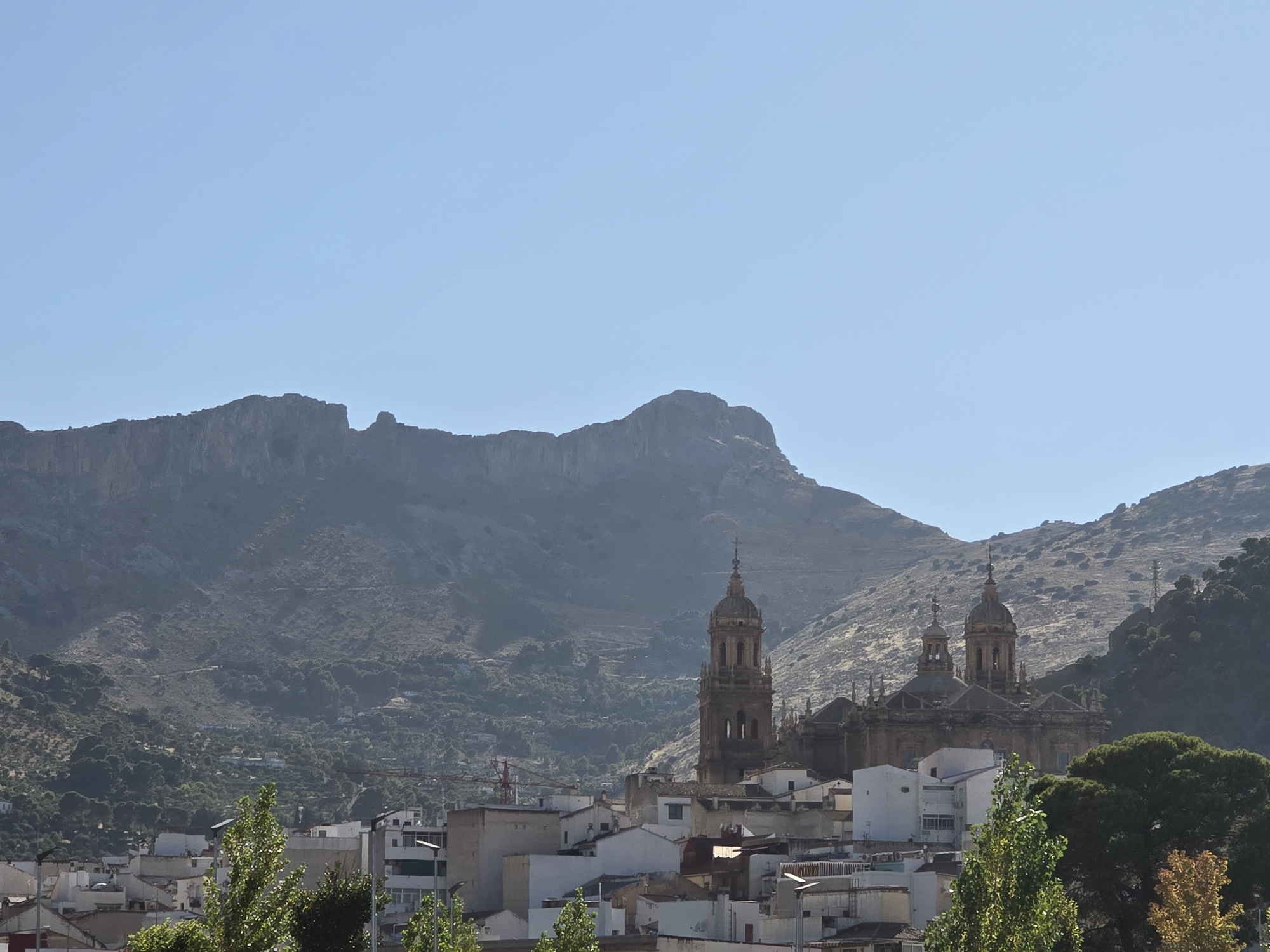 The image shows a cityscape nestled at the base of a large mountain range under a clear blue sky. Dominating the middle ground are two prominent church towers, characteristic of the Cathedral of Jaén. The architecture of the towers, with their intricate detailing and domed tops, suggests a historical and religious significance. Surrounding the churches is a densely packed urban area with white and beige buildings, some with visible windows and balconies.
In the foreground, lush green trees partially obscure the view of the lower buildings, adding a natural element to the scene. The mountain range in the background is rugged and dry, with sparse vegetation, typical of the Andalusian landscape. The overall lighting suggests a bright, sunny day. The photograph appears to be taken from a slightly elevated vantage point, offering a panoramic view of the town and its mountainous backdrop.