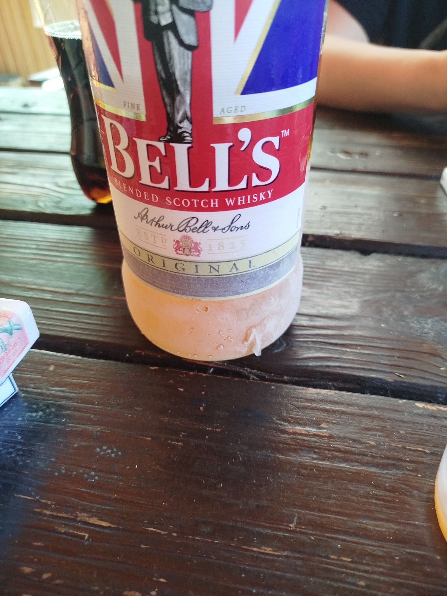 This media file shows a close-up of a bottle of Bell's Blended Scotch Whisky sitting on a dark wooden table. The bottle is mostly red with white lettering and features a picture of a man in formal attire and the Union Jack flag. Text visible on the bottle includes "BELL'S", "BLENDED SCOTCH WHISKY", "Arthur Bell & Sons", "ESTD 1825", and "ORIGINAL". There are water droplets on the lower portion of the bottle, suggesting it is cold. To the left of the bottle, a portion of a cigarette pack is visible, with text that appears to say "SAY", "mod", "B30 26", and "20 WY". In the background, a glass of dark liquid, possibly soda or another beverage, is visible. The scene is outdoors, with hints of a natural light, suggesting it could be daytime. The focus is on the beverage and smoking items, implying a casual setting, possibly a pub or outdoor gathering. The wooden table has a worn texture, with visible grain and some scratches, contributing to a rustic atmosphere.