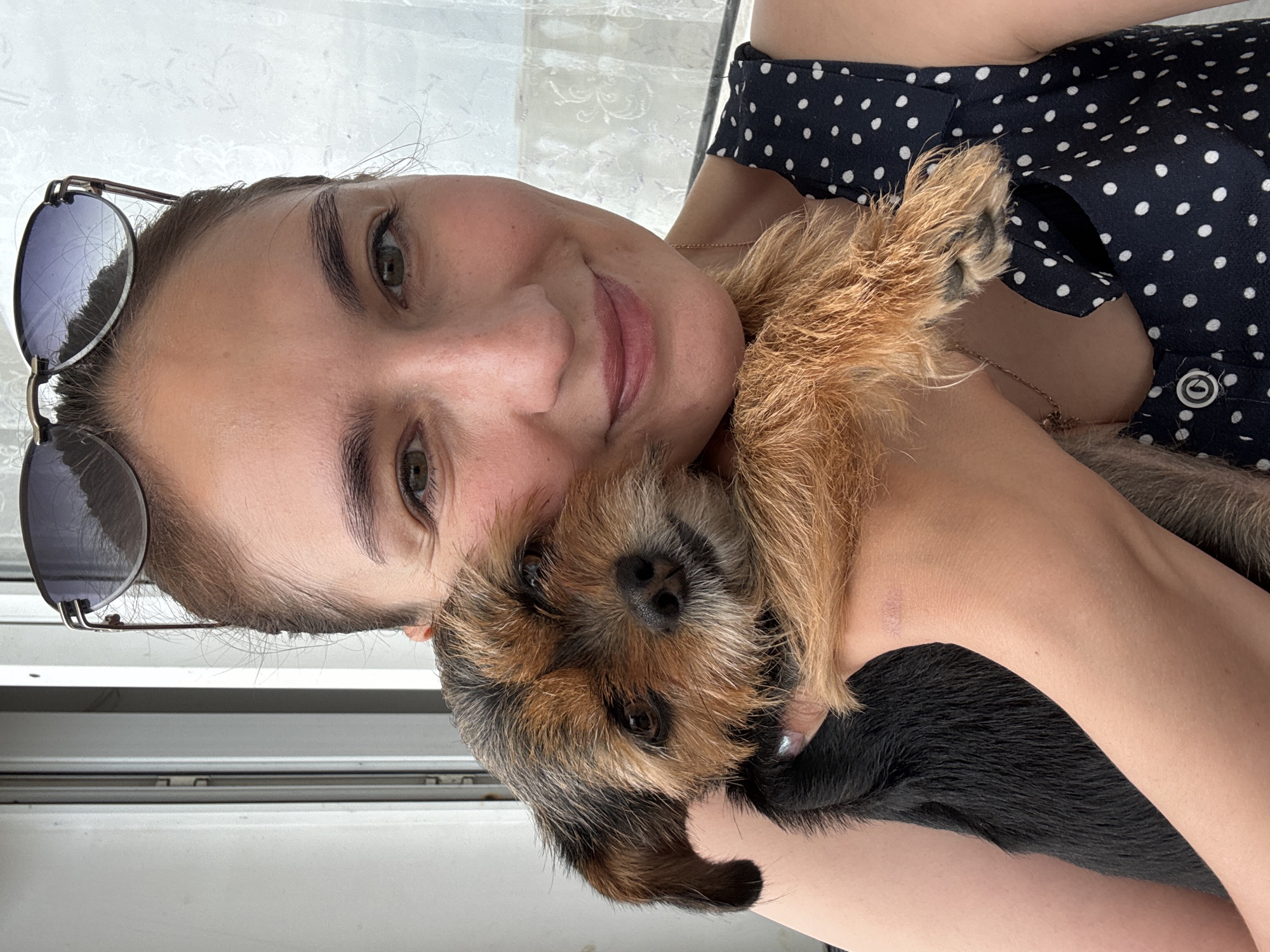 A woman is holding a small, brown and black dog. The woman is wearing a black and white polka-dotted shirt and has sunglasses resting on her head. The dog is nestled close to the woman's chest. The background appears to be a window or balcony with a white frame. The location is not determinable from the information given.