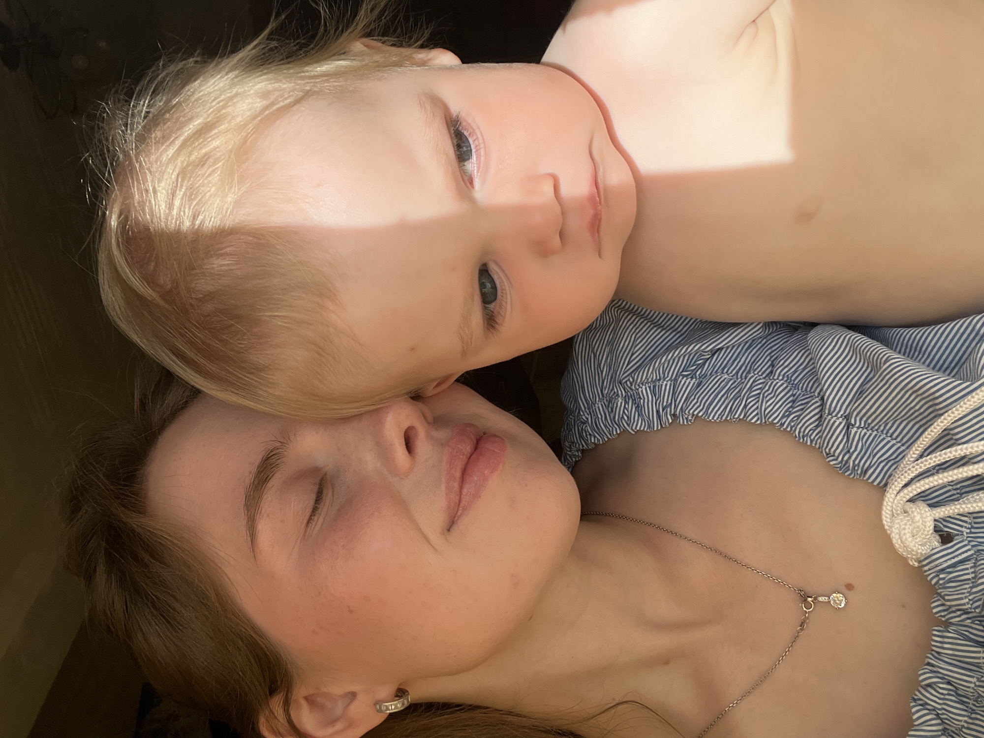 A close-up photograph shows a young child lying on top of a woman. The child has blonde hair and blue eyes, and is looking directly at the camera with a neutral expression. The woman has brown hair, closed eyes, and is lying on her back. She is wearing a blue and white striped shirt and a delicate silver necklace with a small pendant. The setting appears to be indoors, with a warm, natural light illuminating the subjects. The location, Chaikovsky, Russia, cannot be independently verified from the visual information provided.