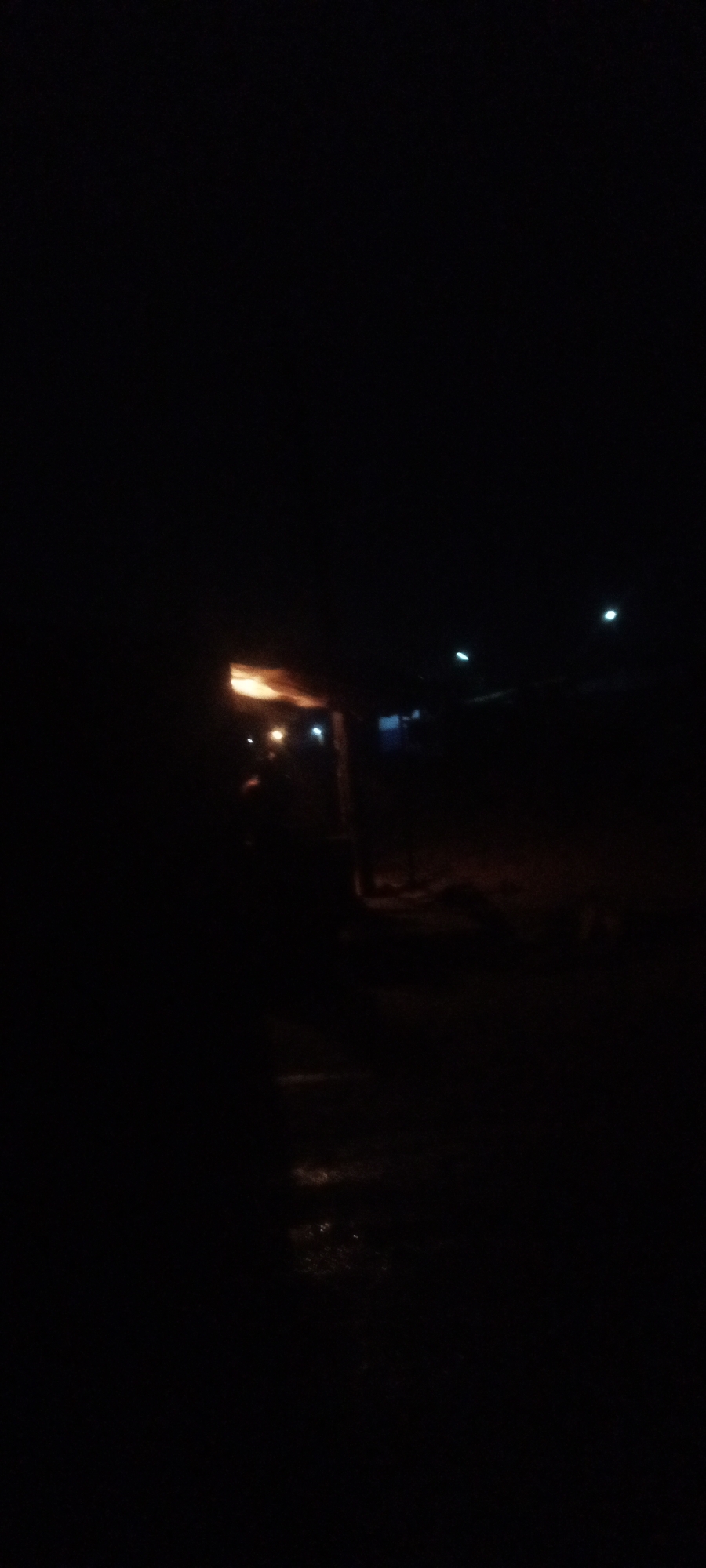 At night in Sabon Tasha, Nigeria, a dark street scene shows a lit building. The scene includes streetlights in the background and a wet road in the foreground. The lighting is poor, with dark shadows obscuring most details.