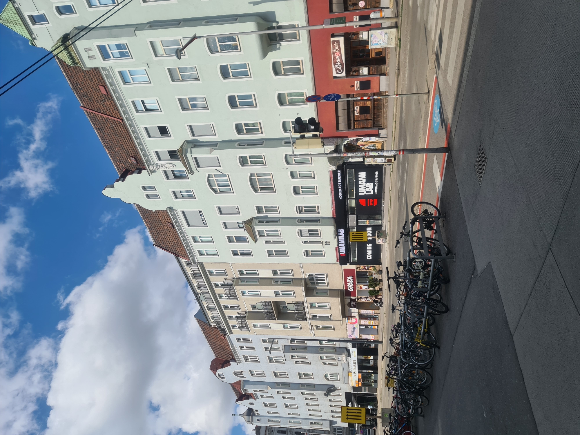 This is a street-level, eye-level, wide-angle shot on a sunny day in Vienna, Austria, looking down a street lined with multi-story buildings. The buildings are a mix of architectural styles and colors, with pastel blue, light grey, and pale green facades. Many of the buildings feature prominent red-tiled roofs and numerous windows. The ground floor of these buildings consists of shops and restaurants, with various signs indicating businesses such as "PARSA MARKET," "GYOZA," "UMAMI LAB HOMEMADE BURGER," and "Brav&Bar." A traffic light is visible on the right side of the frame, along with a crosswalk and a designated bike lane marked with blue paint and a bicycle symbol. A large number of bicycles are parked in racks along the sidewalk in the foreground. There are no people actively engaged in actions within the frame, but a person can be seen walking in the background pushing a stroller. The sky is blue with scattered white clouds.