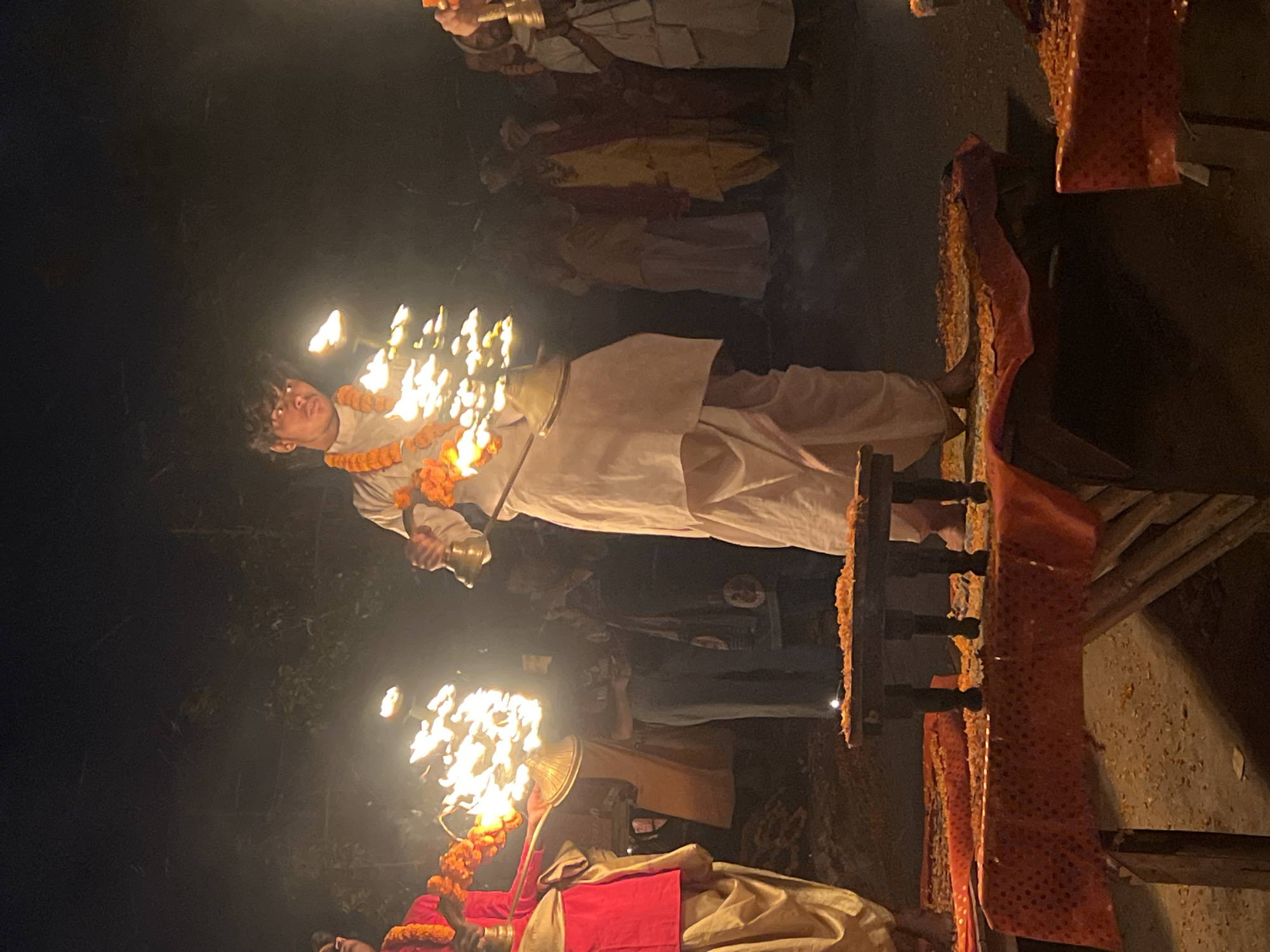 The image shows a religious ceremony, possibly a Ganga Aarti, in Vrindavan, India. A young man, wearing a garland of orange flowers and light-colored traditional Indian attire, stands holding an ornate candelabra with flames. Several other people are also present, some holding similar implements. They stand on a raised platform covered with garlands. The surrounding environment appears to be dimly lit, suggesting it is nighttime. A simple wooden structure with draped fabric is visible in the background.