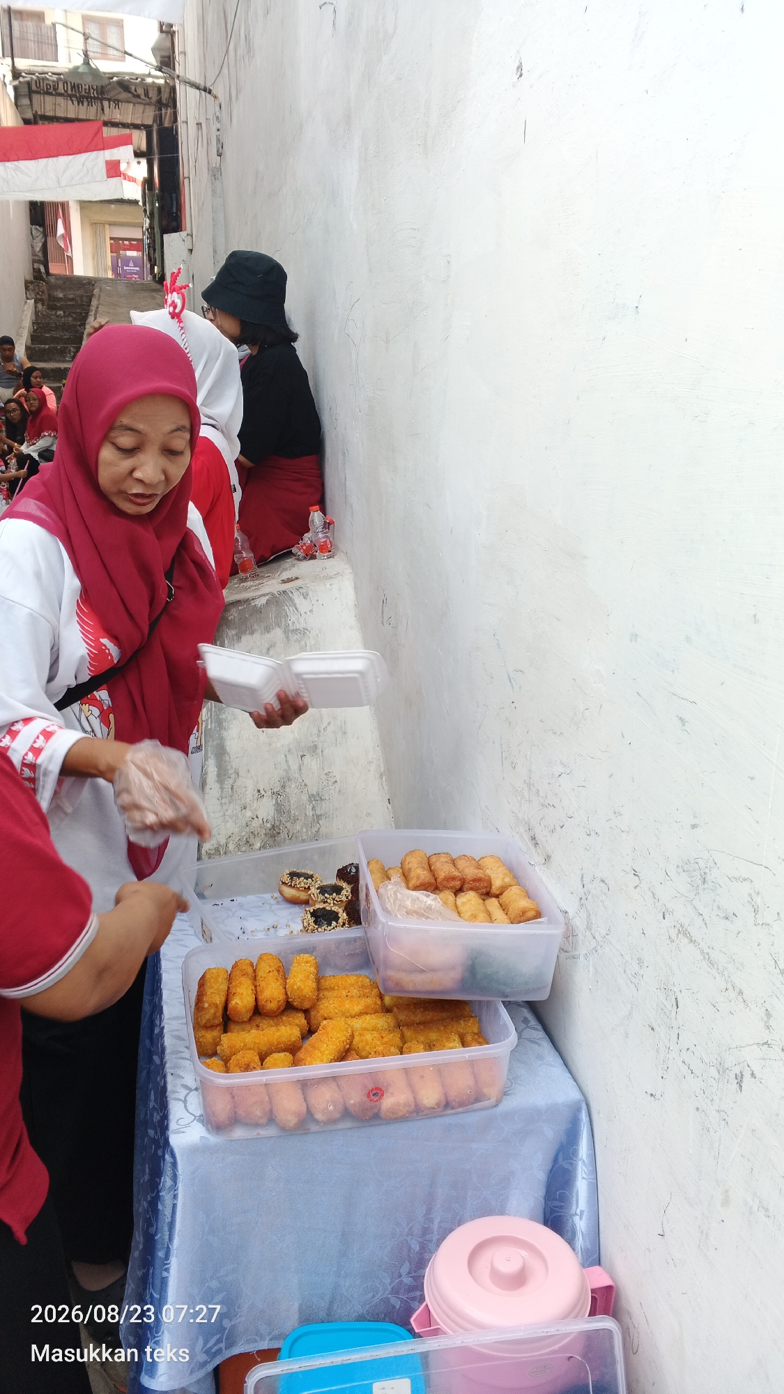 In a narrow alleyway in Malang, Indonesia, a woman wearing a red hijab and a white shirt with red accents is handing food containers to another person. She is standing at a table covered with a blue tablecloth, where several plastic containers of fried food, including what appear to be donuts and spring rolls, are displayed. Behind her, a person in a black hat and red skirt is seated against a white wall. Red and white Indonesian flags are visible in the background, draped over buildings and stairs, suggesting a celebratory or national event. The overall scene depicts a street food vendor operation in a dense urban environment.