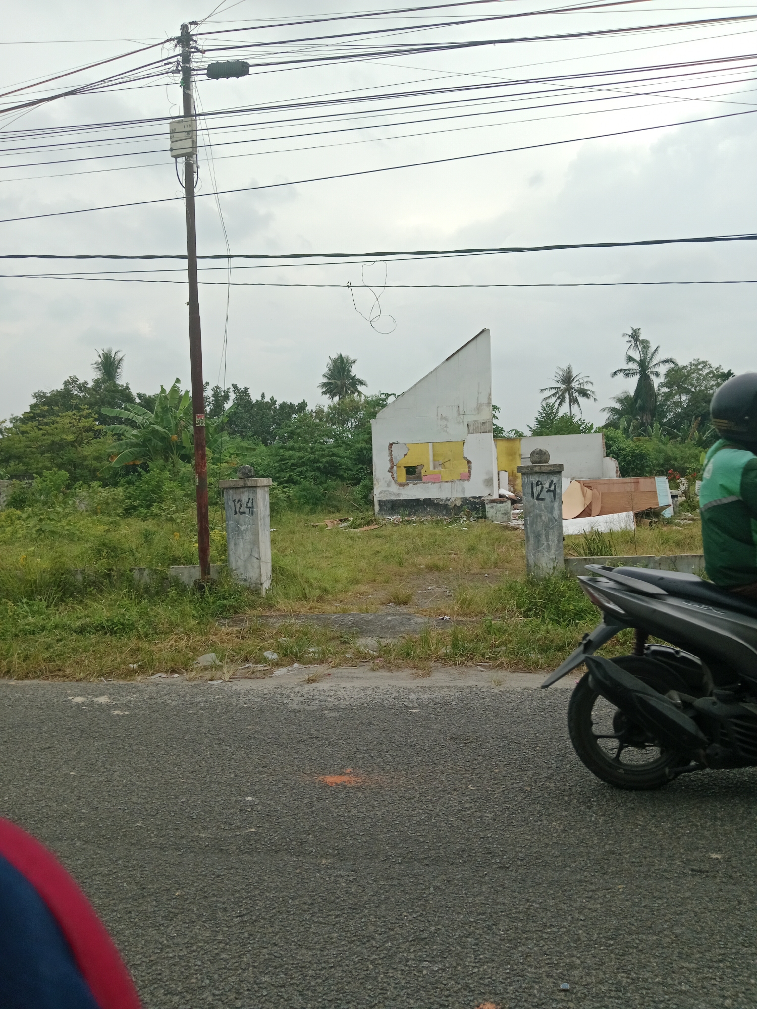 The image shows a street scene in the City of Binjai, Indonesia. A gray asphalt road is in the foreground, with a patch of orange paint visible on it. To the right, the rear of a black motorcycle is parked, with its rider wearing a green and black jacket and a black helmet. Beyond the road, there is a grassy area with a concrete pillar on which the number "124" is painted in black. Further back, a partially demolished white building stands amidst overgrown vegetation and trees, including banana trees and palm trees. Overhead, numerous power lines crisscross the sky, supported by a utility pole. The sky is overcast.