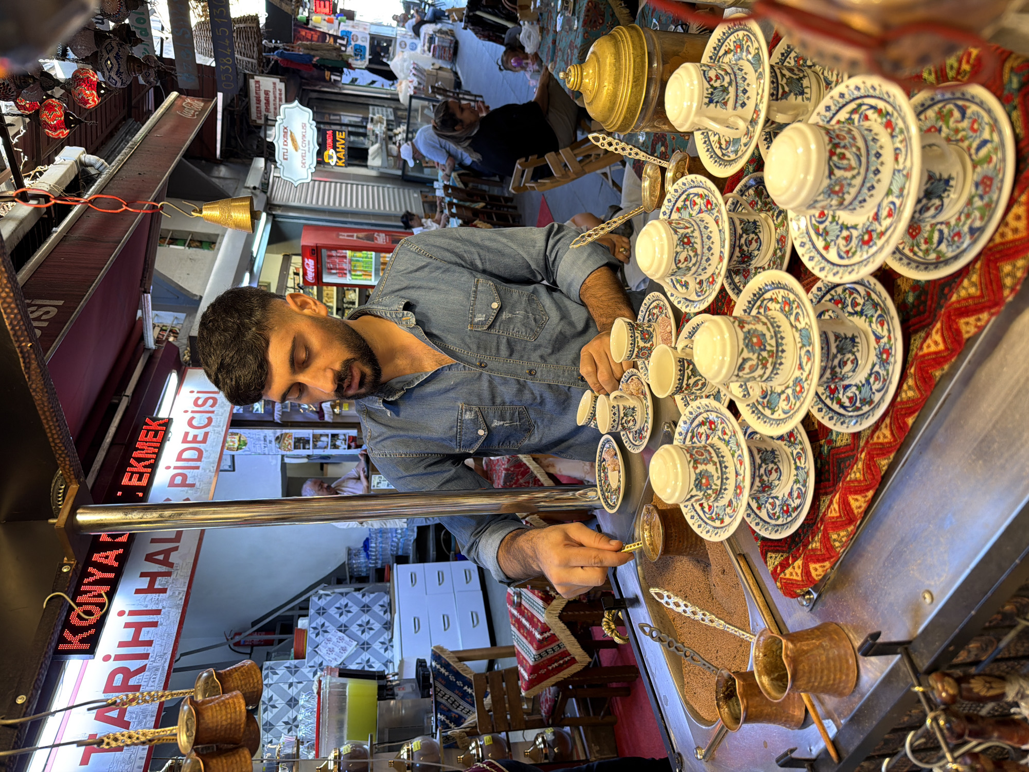In Istanbul, Turkey, a man with dark hair and a beard, wearing a denim shirt, is preparing coffee at a street vendor stall. He is standing behind a metal counter filled with fine sand, where several traditional copper coffee pots (cezves) are placed. In front of the sand, a collection of small, ornate coffee cups and saucers are neatly stacked. The man is holding a small metal spoon, engaged in the process of brewing coffee, with a cezve in the sand. The stall is decorated with a vibrantly patterned textile that is red, yellow, and blue. Behind the man, a sign reads "KONYA TARİHİ HAL" and "PİDECİSİ," indicating a business that specializes in regional food. In the background, other market stalls and people are visible, suggesting a bustling street market environment. A large dispenser of green liquid is on the left, and various decorative items, including lamps, hang from the ceiling.