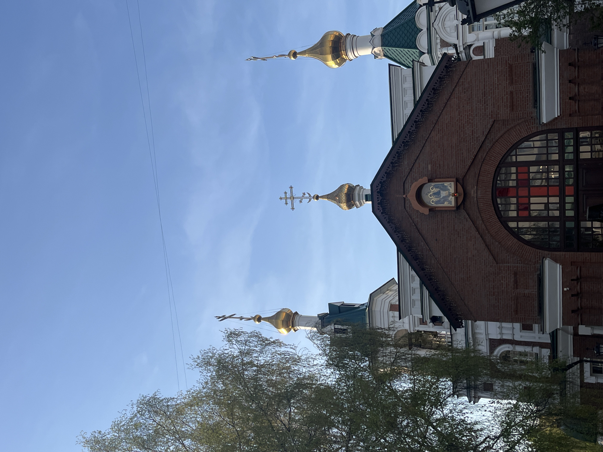 The image shows a view of the Holy Protection Cathedral in Krasnoyarsk, Russia. The building is primarily constructed of red brick with white accents and green roofing. It features multiple golden domes topped with Orthodox crosses. A large, round window is visible on one side of the building, with a smaller icon above it. Tree branches are visible in the foreground. The sky is blue with some wispy clouds. The photo appears to have been taken from a low angle.