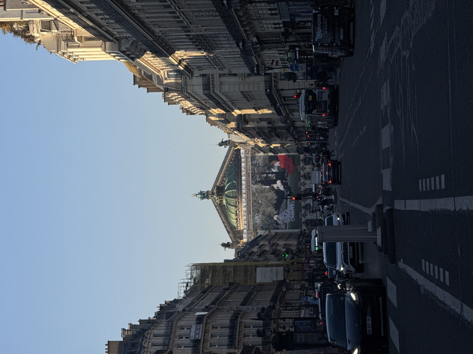 The image displays a street scene on Avenue de l'Opéra in Paris, France, looking southeast towards the Opéra Garnier. The sky is clear blue overhead.
In the mid-ground, the Opéra Garnier stands prominently, characterized by its distinctive green dome and ornate architecture. A large advertising banner, featuring an outdoor scene with three individuals (two in light clothing, one in a red dress) and the "CHANEL" logo, covers the main facade of the opera house.
The street, a multi-lane asphalt thoroughfare, occupies the lower half of the frame and appears dark due to shadows. White painted road markings, including crosswalks and lane dividers, are visible. Traffic is present, with multiple vehicles in motion or stopped. In the foreground left, a dark Tesla Model 3 (license plate JD-622-ZX) faces away from the viewer. Behind it, a white SUV (Toyota RAV4, license plate GL-054-FP) is visible. In the right lane, a city bus and a dark van (with "®loglocons" text) are present. Red and green traffic lights are visible at intersections along the street.
Multi-story Haussmannian-style buildings line both sides of the avenue. The buildings on the right are partially illuminated by sunlight, while those on the left are largely in shadow. Scaffolding covers a portion of a building on the left side. Pedestrians are sparsely visible on the sidewalks and crossing the street in the mid-ground. A white, pillar-like structure is located in the road median in the immediate foreground.