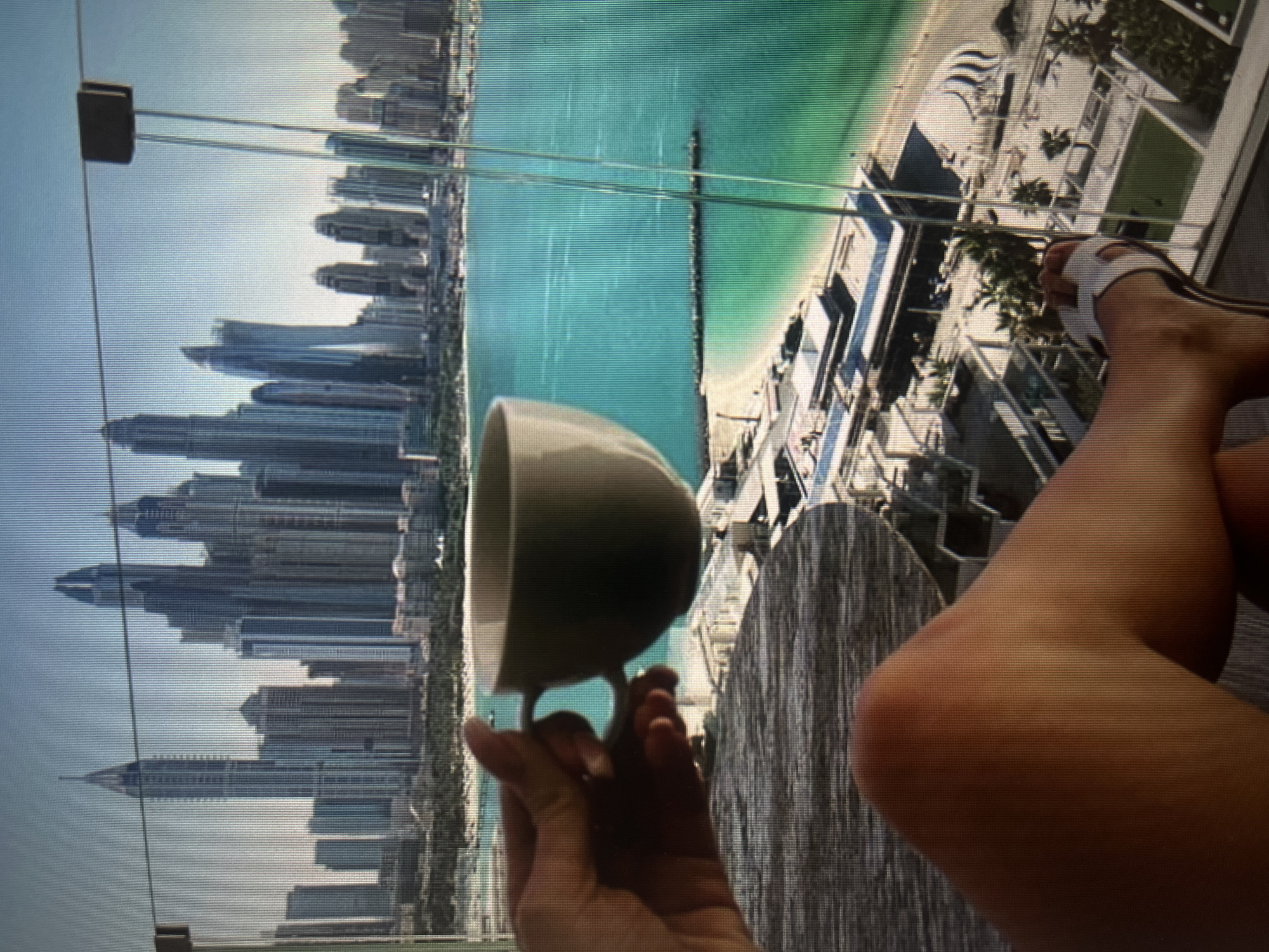 A person's hand is holding a light-colored coffee mug, with the thumb and forefinger gripping the handle, against a backdrop of a city skyline and ocean. The mug is positioned in the foreground, obscuring part of the view. In the mid-ground, a turquoise body of water is visible, with a sandy beach and a breakwater extending into the sea. Beyond the water, a dense collection of modern skyscrapers dominates the skyline, characterized by varied architectural styles and heights. The sky above is a clear, pale blue. A person's leg, clad in a white sandal, is visible in the lower right portion of the image, suggesting the perspective is from a balcony or elevated vantage point. The overall scene is a snapshot of urban life combined with a coastal environment, suggesting a vacation or a scenic residential area. The location is not Moscow, Russia.