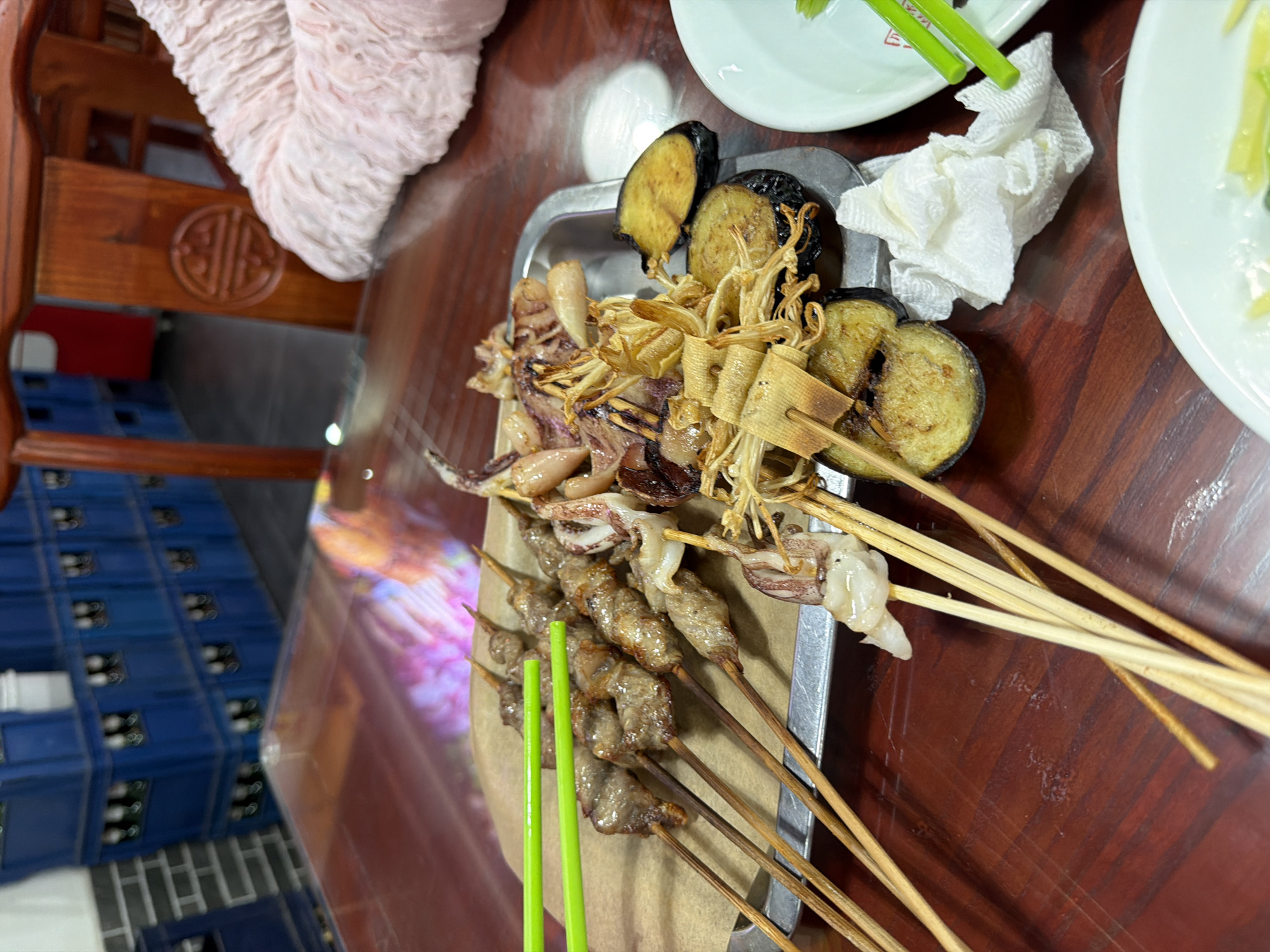 Captured from an elevated perspective, the image shows a close-up of a metal tray on a wooden surface, presumably a table. The tray holds a variety of skewered foods: grilled squid, tofu skins, eggplant, and other types of skewers. The wooden surface reflects light. Green chopsticks lie on the tray. Next to the tray, a white tissue sits. In the background, partly visible, is a wooden chair, and stacked blue boxes, possibly beer crates, are also noticeable. The image was taken indoors, likely in the Dongcheng District of China.