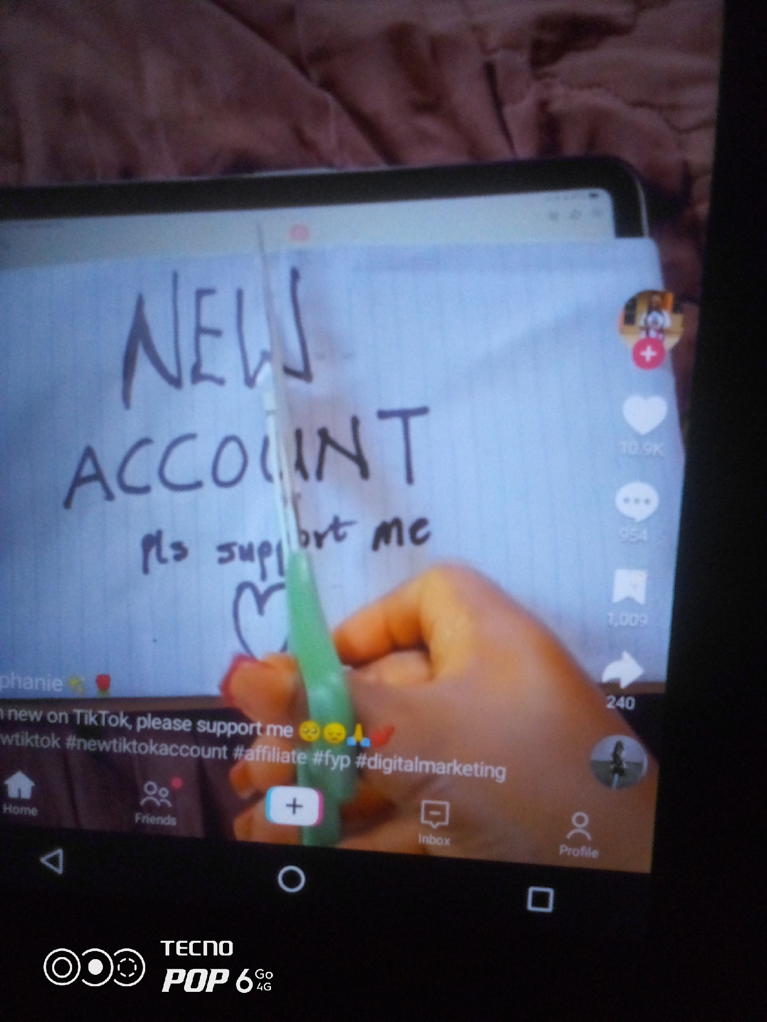 The image depicts a close-up view of a tablet screen displaying a TikTok video. On the screen, a handwritten sign on white paper reads "NEW ACCOUNT pls support me." Below the text, there's a drawn heart and a finger holding a green stylus or pen. The video interface elements of TikTok are visible, including like, comment, share, and profile icons with counts. A hand with orange-toned skin and red-painted fingernails is interacting with the screen. The background of the image is a blurred reddish-brown fabric, possibly a blanket or bedspread. The bottom of the image shows a smartphone interface with navigation buttons and the "TECNO POP 6 Go 4G" branding. The scene does not provide any information about the city of Howayi, Nigeria.