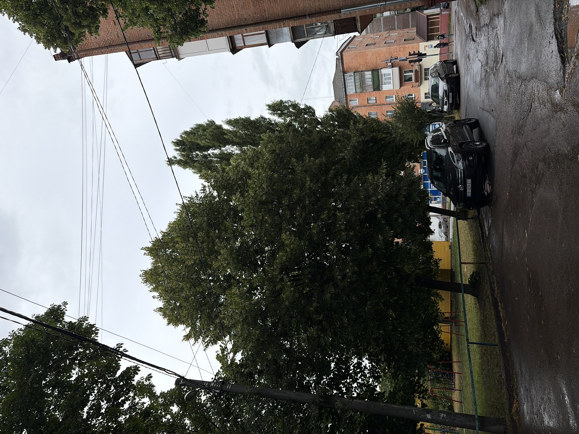 The image depicts a street scene in Chernihiv, Ukraine, under an overcast sky. In the foreground, a wet asphalt road with potholes is visible. To the left, a playground with yellow fencing and red swing sets is partially obscured by trees. A dark-colored Volkswagen SUV, license plate CB 4234 ET, is parked on the street. Further down the street, another dark-colored car, a Skoda station wagon, license plate CB 7268 ET, is also parked. Two individuals, a male and a female, are walking away from the camera on the right side of the road. Behind the parked cars and pedestrians, there are multi-story brick buildings with balconies and windows. Overhead, numerous electrical wires are strung between poles and buildings. The overall environment suggests an urban residential area.