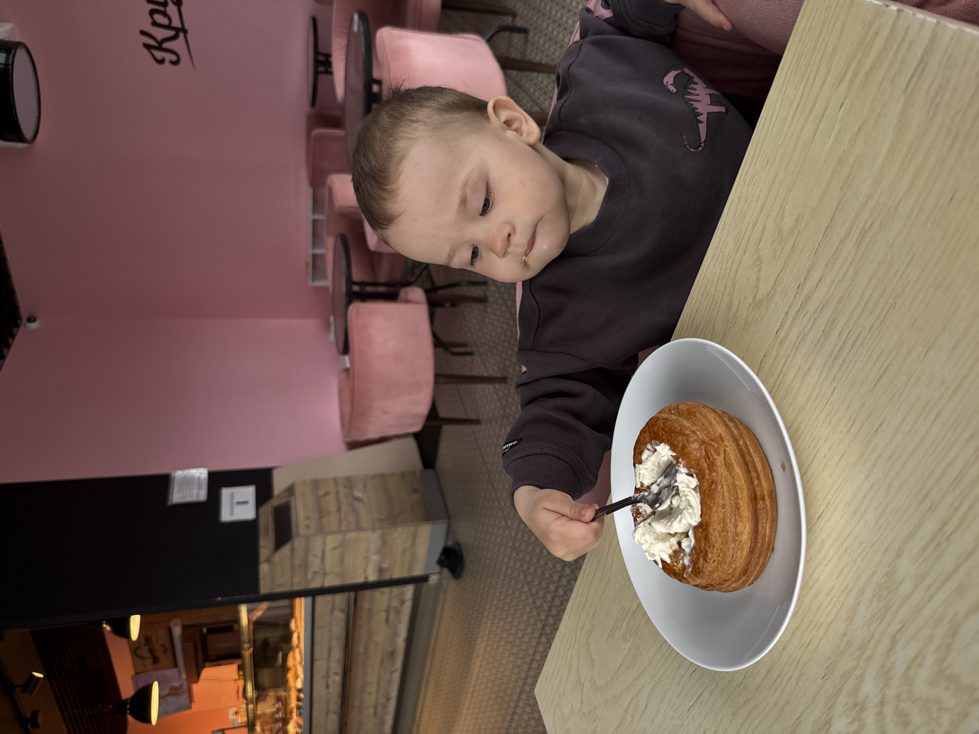 The image displays a young child engrossed in eating a pastry within a cafe setting, likely located in Rusanovo, Russia. The child, a toddler with light brown hair, is wearing a dark grey long-sleeved top featuring a pink dinosaur-like graphic on the left arm. They are seated at a light-colored wooden table, their attention focused on a large, golden-brown, round pastry, possibly a cruffin or large bun, presented on a white oval plate. The pastry is generously topped with white whipped cream, into which a spoon is inserted. A small smudge of cream is visible on the child's chin.
The background reveals a cafe interior with distinctive decor. A prominent pink wall features dark, cursive lettering, partially visible as "Кри" or "Кру". Numerous pink upholstered chairs with dark metal frames are arranged around small tables, suggesting a welcoming seating area. A darker wall section displays some papers and a white square sign with the number "1" clearly marked. The floor appears to be carpeted with a textured, light brown material. Reflections in a large mirror or glass partition on the left side of the frame show additional interior elements, including wooden structures and lighting, contributing to the cozy ambiance. The child appears calm and concentrated on their treat.