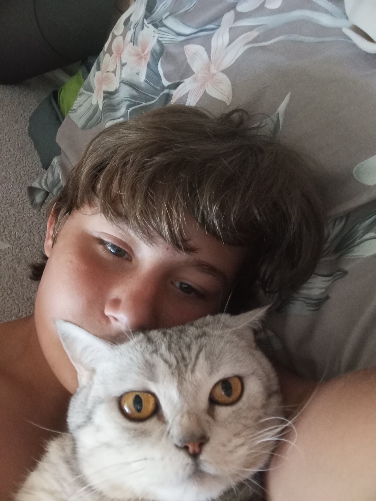 The image is a close-up selfie of a young person and a cat, taken from a low angle. The person, with short brown hair and a light complexion, is lying down, with their head resting on a pillow that has a floral pattern in shades of pink and gray. Their left eye is open and looking towards the camera, while their right eye is partially obscured. A gray and white cat with striking amber eyes is positioned in front of the person's face, its head largely filling the lower portion of the frame. The cat's fur appears soft and textured. The background consists of a textured surface, likely bedding or a blanket, and a hint of a dark object on the left, possibly furniture. The overall impression is one of intimacy and comfort, suggesting a domestic setting. The image does not contain any visual elements that would indicate its location in Sozonivka, Ukraine.