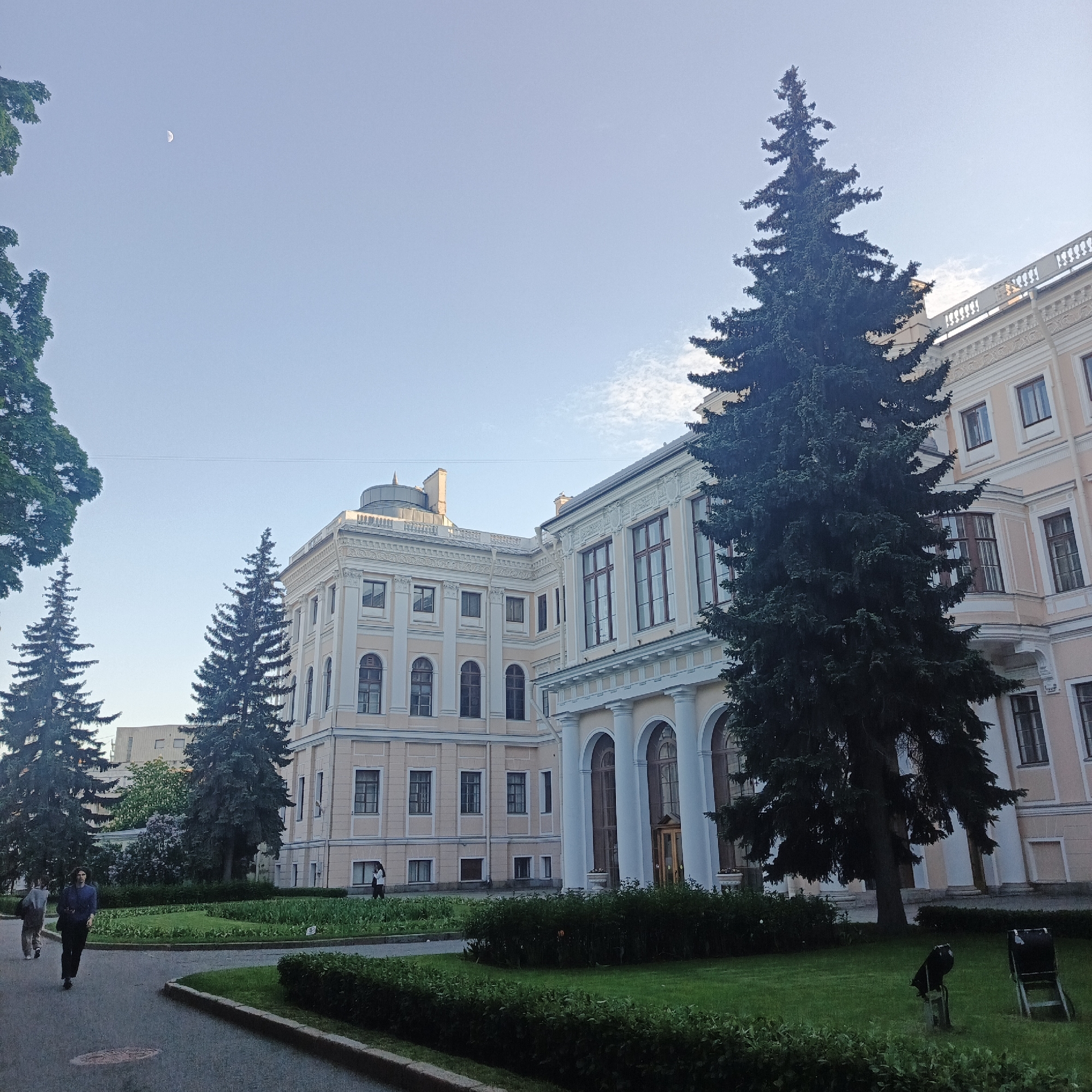 The image depicts a view of the Mikhailovsky Palace in Saint Petersburg, Russia. The palace is a large, ornate building with a pale yellow facade, white columns, and numerous windows. Several tall, dark green spruce trees are planted in front of the palace, partially obscuring the view. There is a well-maintained lawn and manicured hedges in the foreground.
Two people are walking along a paved path in the lower left of the frame. In the sky, a thin crescent moon is visible in the upper left corner. The sky is a pale blue with a few scattered clouds.
