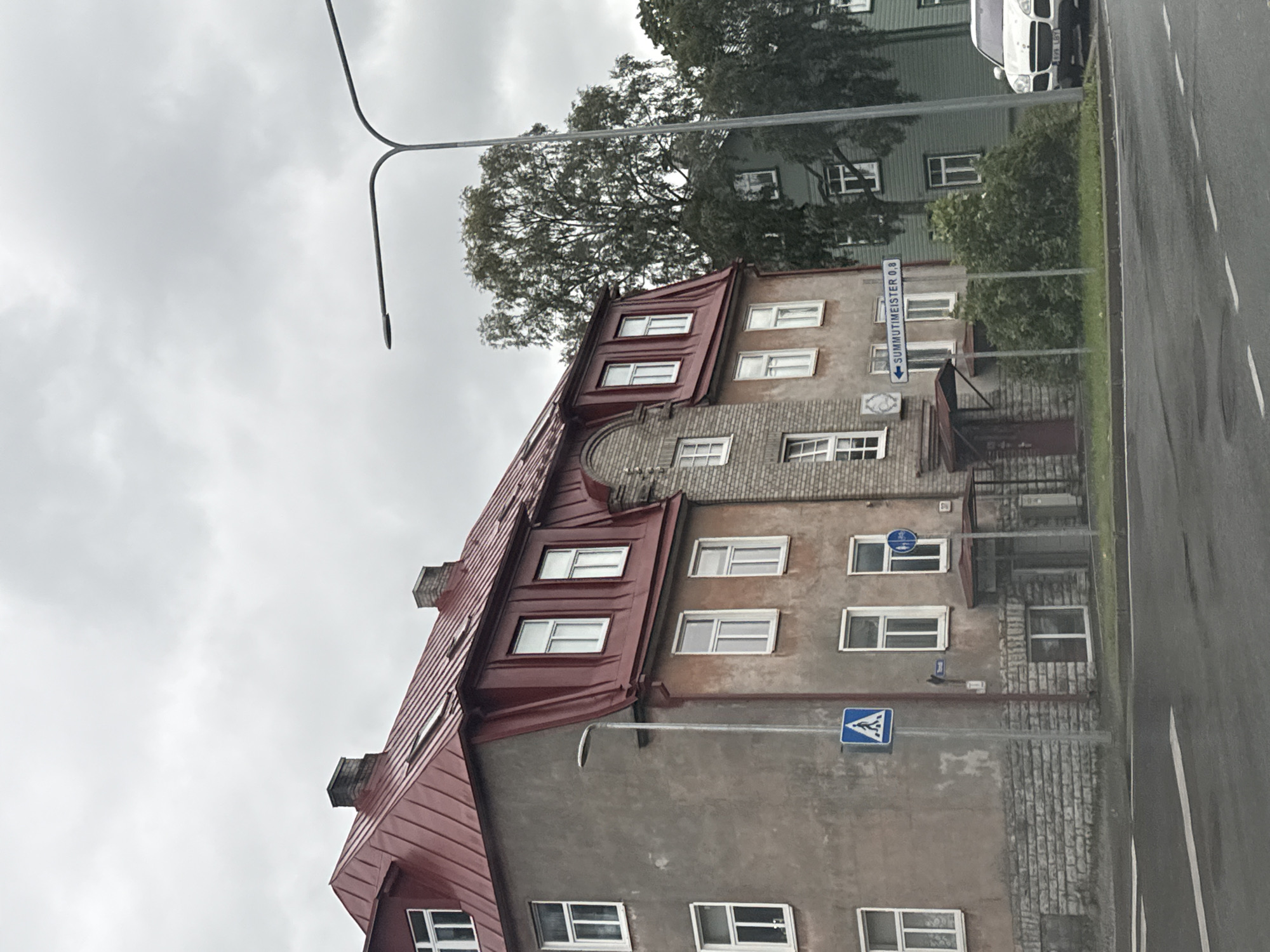 The image captures a street view in Tallinn, Estonia, under an overcast sky. In the foreground, a paved road with white lane markings runs across the bottom of the frame. To the right of the road, a white SUV is parked.
Dominating the left and center of the image is a multi-story building with a distinctive red, sloped roof. The building's facade is a mix of textured plaster and brickwork. Several windows with white frames are visible on its various levels. A tall lamppost stands near the building, with a blue square sign featuring a white pedestrian symbol mounted on it. Further to the right, a street sign with the text "SUMMUTIMEISTER 0,8" and an arrow pointing left is visible. Behind the main building, a green building with similar windows can be seen, partially obscured by trees. The overall atmosphere is muted, consistent with the cloudy weather.