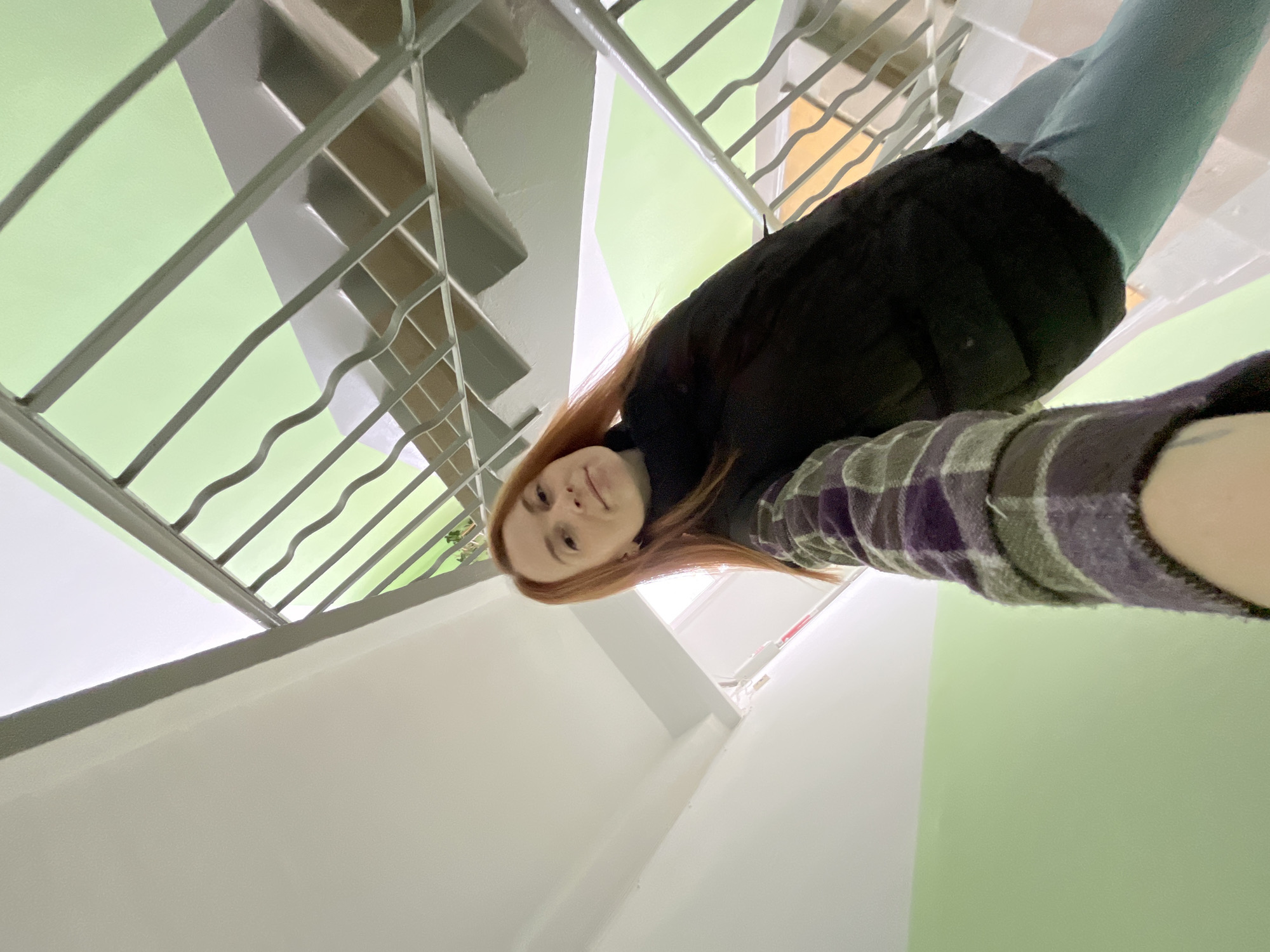 A young woman with red hair is taking a selfie inside a stairwell of a building. She's wearing a dark jacket and a plaid shirt. The stairwell has white walls and green painted sections. A metal railing is visible along the stairs. The exact location in Chaikovsky, Russia cannot be determined from the visual information.