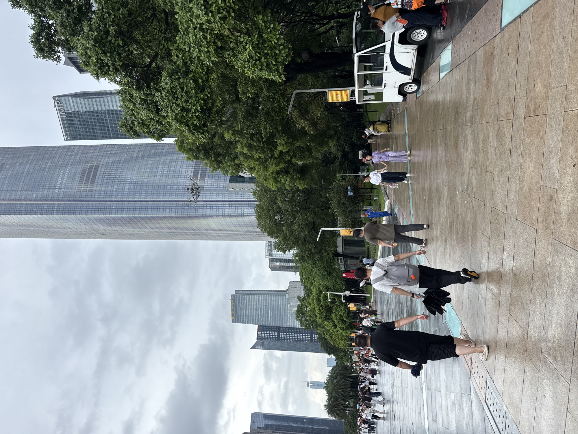 This is an outdoor scene in Tianhe District, China, on a cloudy day. The pavement is wet, indicating recent rain. In the foreground, two men are walking away from the camera. The man on the left wears a black t-shirt and black shorts, carrying a folded black umbrella. The man on the right wears a white t-shirt, black pants, and a grey backpack, also carrying a folded black umbrella.
Further down the paved walkway, a group of people are walking in various directions. To the right, a white electric vehicle is parked, with several people standing near it. In the background, several modern skyscrapers dominate the skyline, some with glass facades. Lush green trees line the walkway on the right side, interspersed with streetlights and signage. The sky is overcast with heavy clouds.