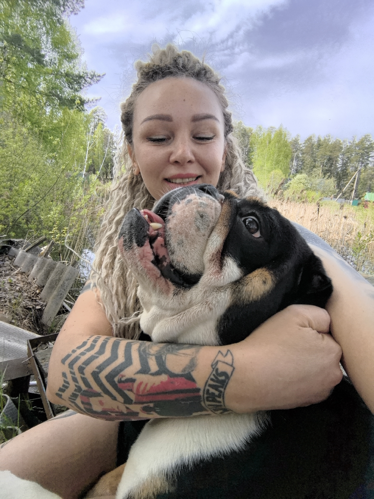 A fair-skinned woman with blonde dreadlocks smiles down at a black, white, and tan English Bulldog dog in her arms. The woman is wearing what looks like a sleeveless shirt, and has tattoos covering both arms. The dog is looking up to the left of the woman with its mouth open. In the background is a rural landscape with trees, tall grass, and a small house. Weather appears to be overcast. Based on location, it is likely in Noviy, Russia.