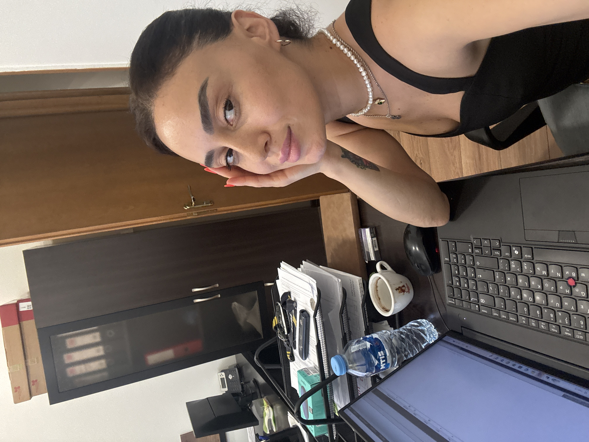 A woman with dark hair and tan skin is sitting at a desk in what appears to be an office. She is looking at the camera and resting her head on her hand. She is wearing a black top and a pearl necklace. On the desk in front of her is a laptop, a water bottle, a coffee mug, and a computer mouse. In the background, there is a tall cabinet with boxes stacked on top of it, and a door. The room appears to be well-lit. The scene is indoors. The image is a selfie.