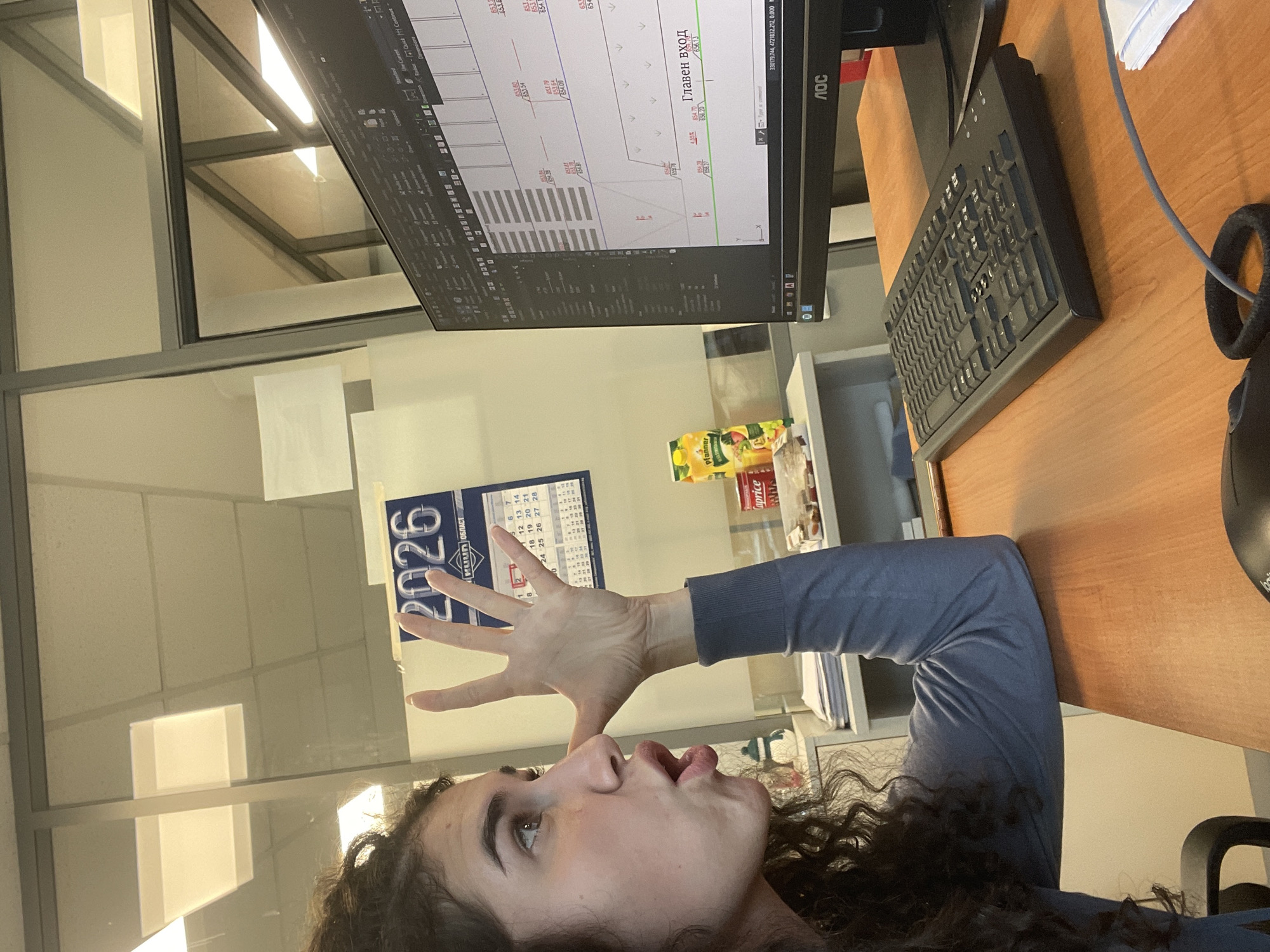 The image captures a candid moment of a young woman in an office environment. She is shown in profile from the chest up, with dark, curly hair, looking upwards and slightly to the right with her mouth agape in an expression of surprise or animated speech. Her left hand is raised, fingers splayed, as if gesturing or emphasizing a point. She is wearing a blue-grey long-sleeved top.
She is seated at a wooden desk, which holds a black AOC computer monitor displaying what appears to be a CAD or architectural drawing. The screen shows lines, numerical data like "654.70", "655.79", and "654.13", and Cyrillic text including "Главен вход" (Main entrance). A black computer keyboard is on the desk, along with a black headset or headphones visible on the right. Several beverage containers are also present, including a yellow Pfanner juice carton and a red Caprice can.
Behind the woman, a wall calendar for the year "2026" is prominently displayed, showing a grid of days and the Cyrillic word "ОБЛАСТ" (District/Region). The background features bright overhead lighting reflected in glass partitions, indicating a modern office setting. The presence of Cyrillic text and the office environment align with the location context of Sofia, Bulgaria. The image portrays a dynamic, informal moment, likely during a workday.