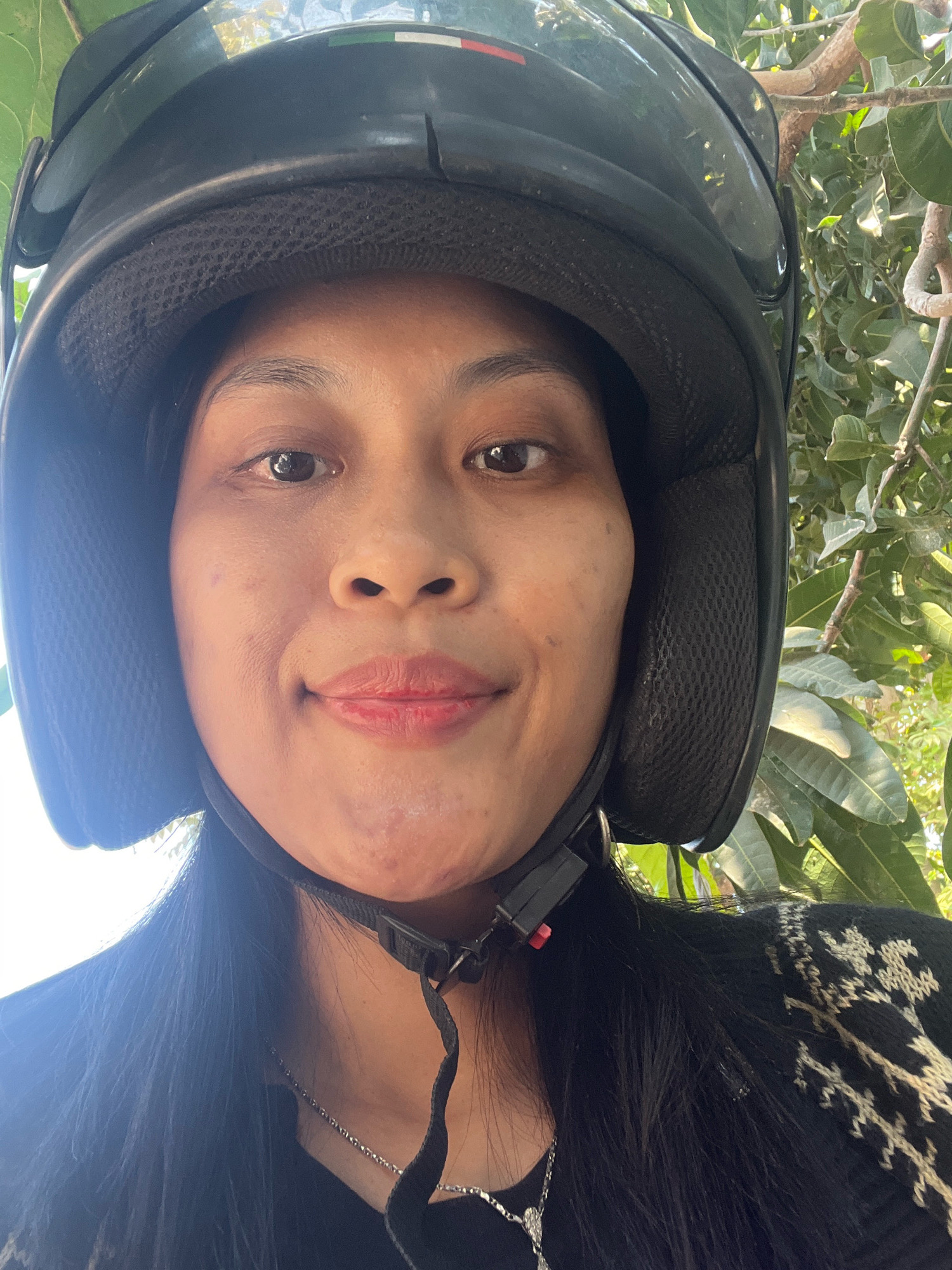 A close-up selfie shows a person wearing a black helmet and a black sweater with a white pattern. The person has brown eyes, a small nose, and red lipstick. The background is blurred with green foliage, suggesting an outdoor setting. The image appears to have been taken in Kecamatan Pati, Indonesia, as indicated by the location data. The overall scene is casual and personal, likely captured during a motorcycle ride or a break.