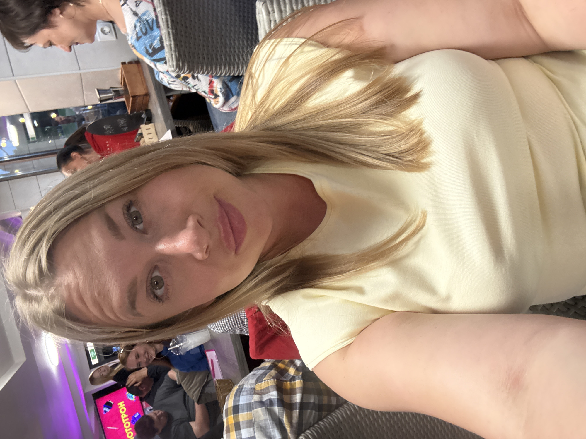 A medium shot selfie features a blonde woman in a light yellow top, her face angled towards the camera. She has green eyes and is looking into the lens. In the background, other people are visible in what appears to be a restaurant or cafe. To the left, a screen with pink and purple lighting displays text, possibly advertisements. Further in the background, individuals are seated at tables, some engaging in conversation. A woman with dark hair and hoop earrings is seen in profile on the right side of the frame. The setting suggests an indoor public space, possibly a restaurant in Veliky Novgorod, Russia, given the signage and overall ambiance.