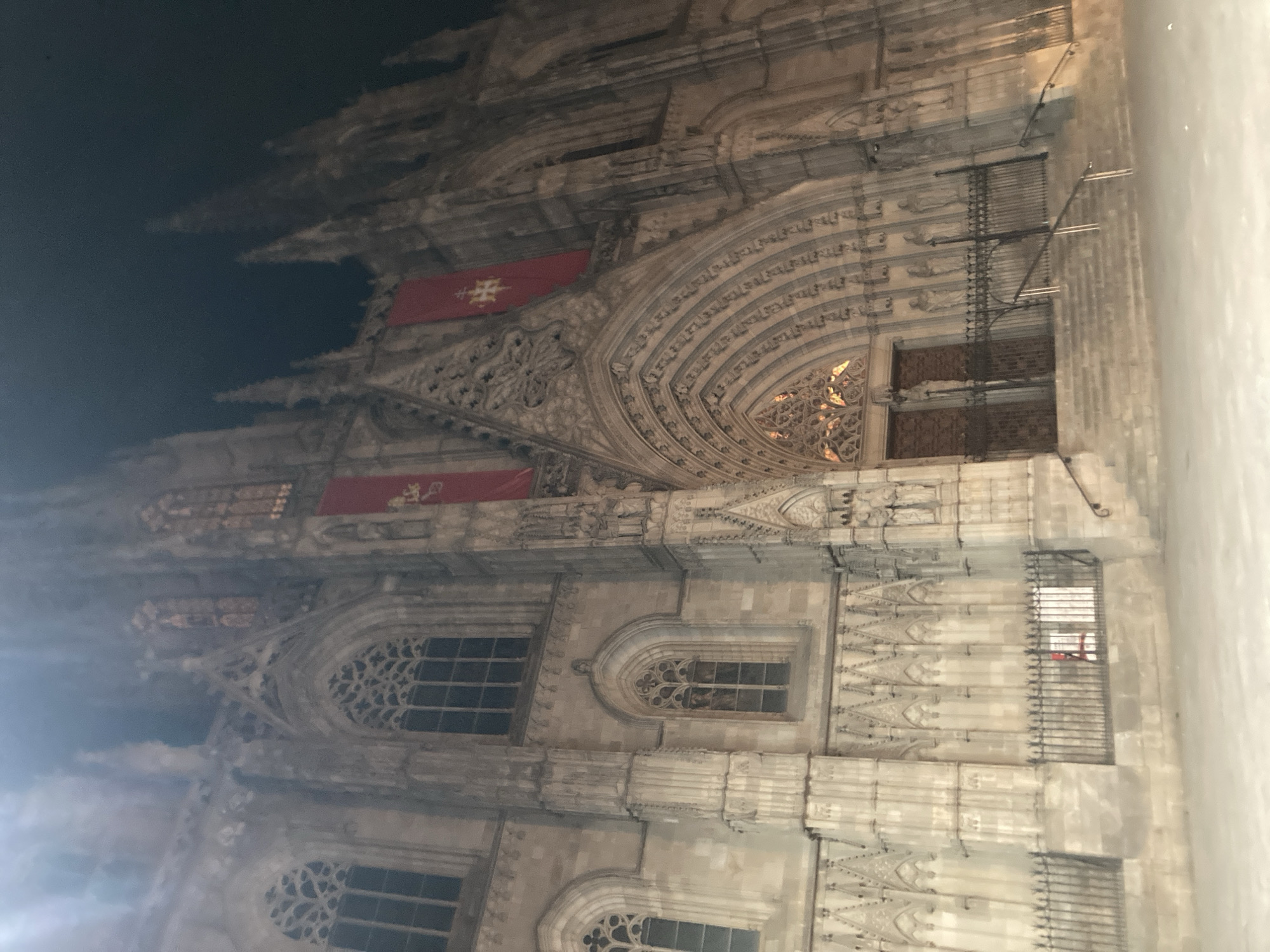 The image captures an exterior night view of the Barcelona Cathedral (Cathedral of the Holy Cross and Saint Eulalia), located in the Gothic Quarter of Barcelona, Spain. The grand Gothic facade of the cathedral is illuminated from below, highlighting its intricate architectural details against a dark night sky.
The central feature is the main portal, deeply recessed with multiple sculpted archivolts and statues in niches on either side. Dark double doors are visible within the portal, secured by a decorative wrought-iron gate at the base of the stairs. Above and to the sides of the central portal, two large rectangular red banners, each adorned with a golden patriarchal cross emblem, hang prominently.
The facade displays numerous Gothic elements, including pointed arch windows with elaborate tracery, such as a large rose window on the upper left side and several lancet windows below. Pinnacles and smaller spires rise towards the top of the building. The lower part of the facade features a detailed stone balustrade and a black wrought-iron fence running along the base, behind which indistinct white signs are partially visible. A wide set of stone steps with metal handrails leads up to the main entrance. No individuals are present in the scene. The lighting creates a dramatic effect, emphasizing the texture and depth of the ancient stone structure.