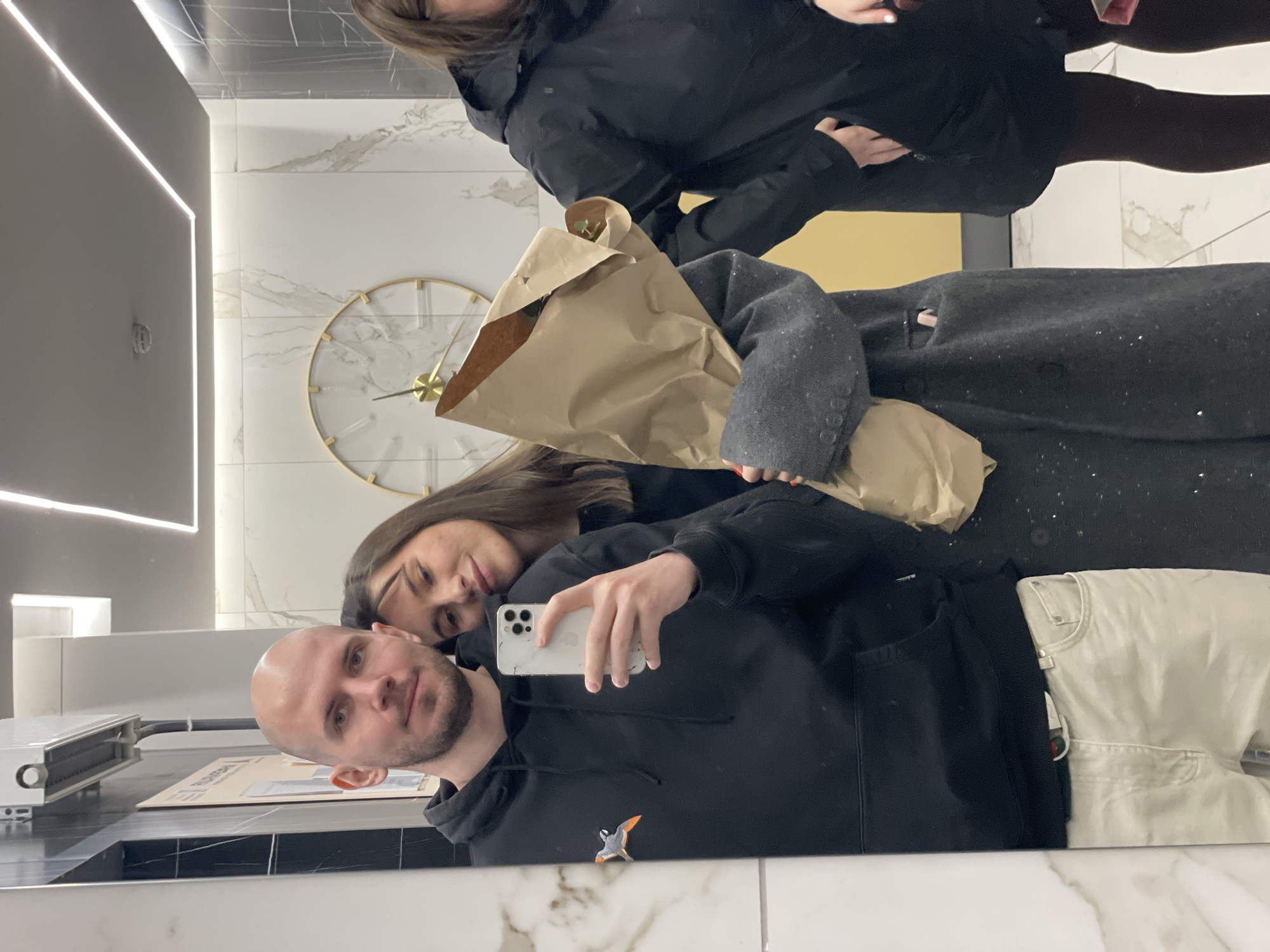 A photograph captures a couple in what appears to be a public restroom setting. The man is bald with a beard, wearing a black hooded sweatshirt, and holding a smartphone, possibly taking a selfie. The woman is leaning in close to him, holding a brown paper bag that appears to contain flowers or some other gift. Another woman, partially visible, is positioned above them.
The background features marble-like wall tiles and a large, decorative clock. There's also a metal fixture and other elements indicative of a restroom environment, such as a mirror and a radiator.
Given the limited contextual information, determining the precise location within Krasnoyarsk, Russia, is difficult. Without further clues, it would be impossible to verify.