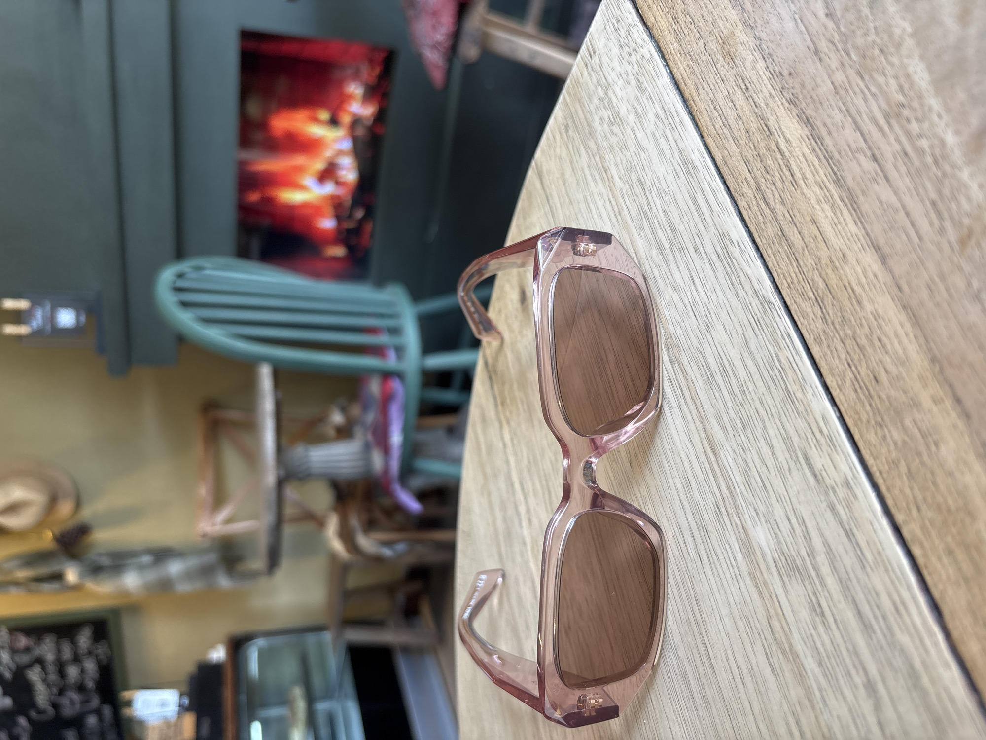 The image shows a pair of pink-framed sunglasses resting on a light brown wooden table, possibly in an indoor setting. In the blurred background, there's a green chair, a display on a wall, and other indistinct elements suggesting a cafe or restaurant environment. Given the request for a location in Kuta Selatan, Indonesia, the image might depict sunglasses left on a table in a cafe or eatery within that area. No people are visible in the scene.