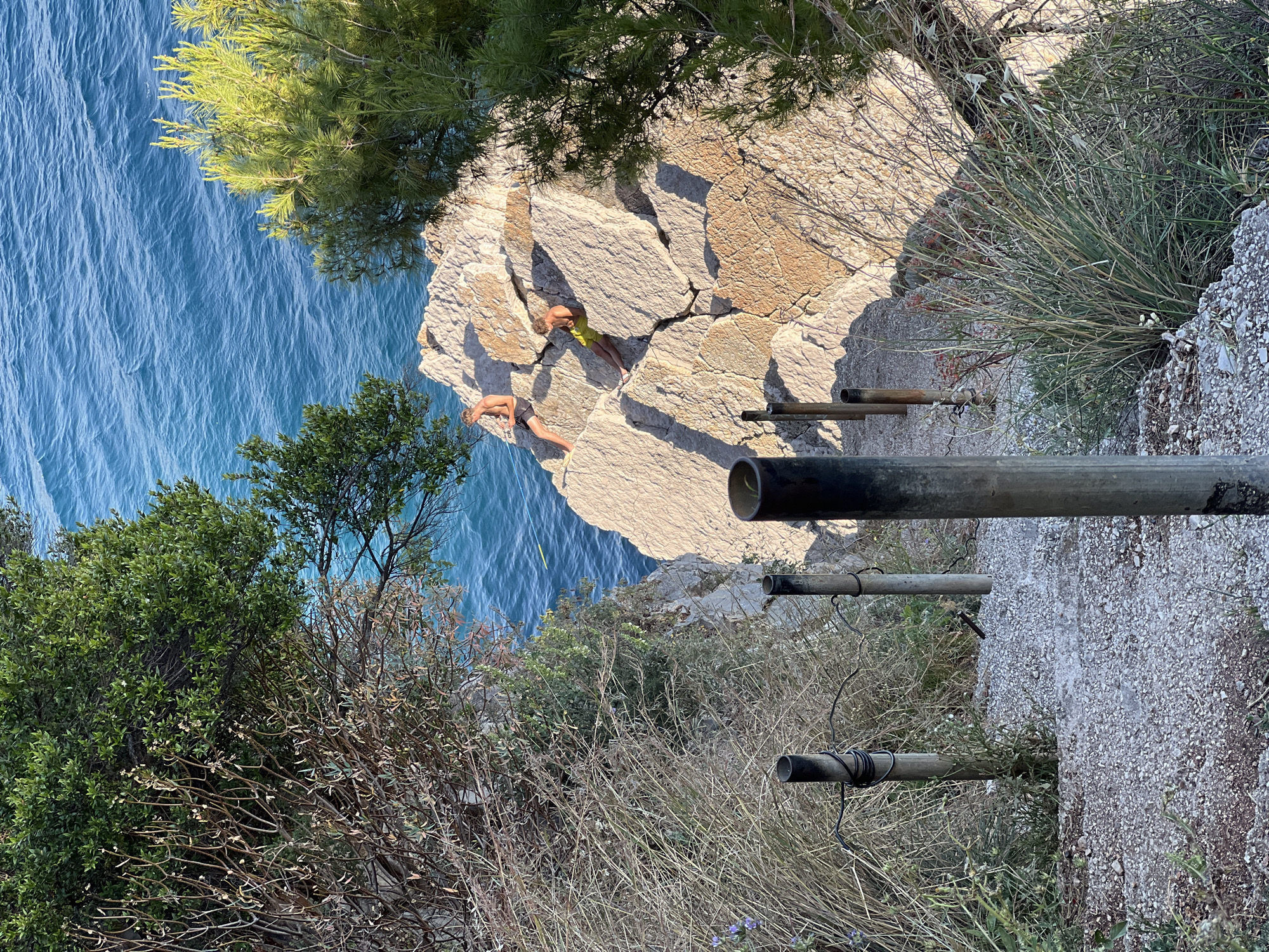 The image shows a rocky coastline in Medinski Krš, Montenegro. Turquoise water can be seen below a rocky cliff. Two people lie prone on the cliff's edge. A series of metal pipes are attached to a steep path leading away from the cliff. Vegetation including trees, shrubs, and grasses is visible. The scene appears to be taken from a high vantage point.