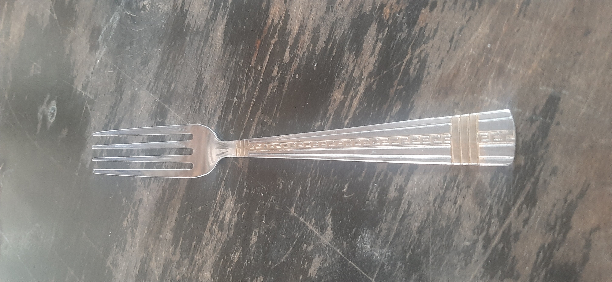 A single, four-tined metal fork lies horizontally on a dark, wood-grain surface. The fork is positioned vertically in the image, with the tines pointing towards the top edge. The handle of the fork has a decorative pattern featuring horizontal bands and a textured, ridged design. The background appears to be a table with a distressed, dark wood finish. There are no people visible in the image, and no discernible actions or interactions are taking place. The scene is static and appears to be an indoor setting. The location is Yessentukskaya, Russia.