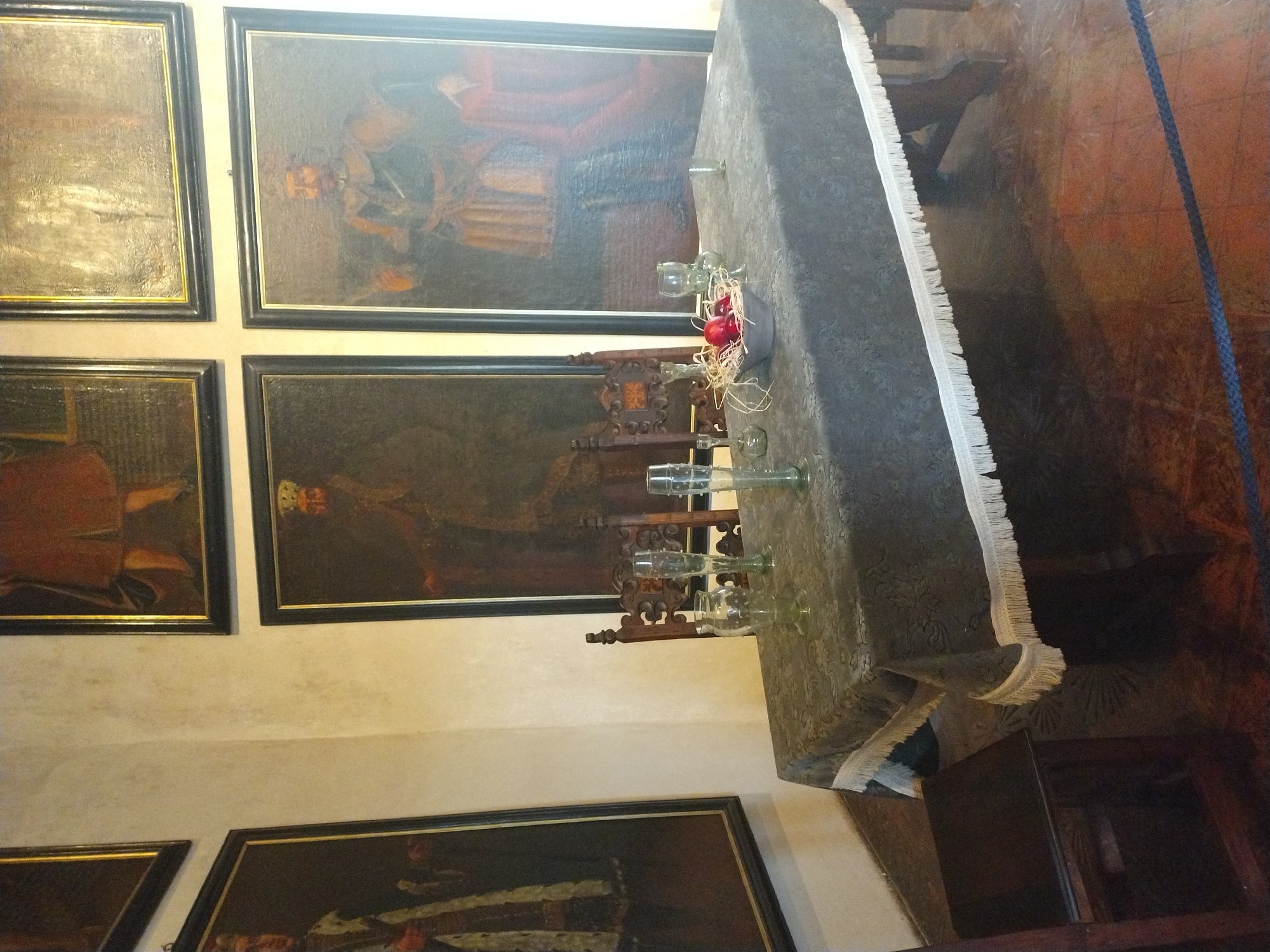 The image shows a room with several large framed paintings on the walls, depicting what appear to be portraits of historical figures. In the center of the room, a rectangular table is covered with a dark tablecloth that has a fringe trim. On the table, there are several glass objects, including a vase and drinking glasses, as well as a bowl containing red fruit, possibly apples, surrounded by straw. Two wooden chairs with ornate backs are positioned behind the table. To the left, a wooden chair is partially visible. The floor is made of wooden planks. This scene is located in the Great Hall of Karlštejn Castle in Karlštejn, Czechia.