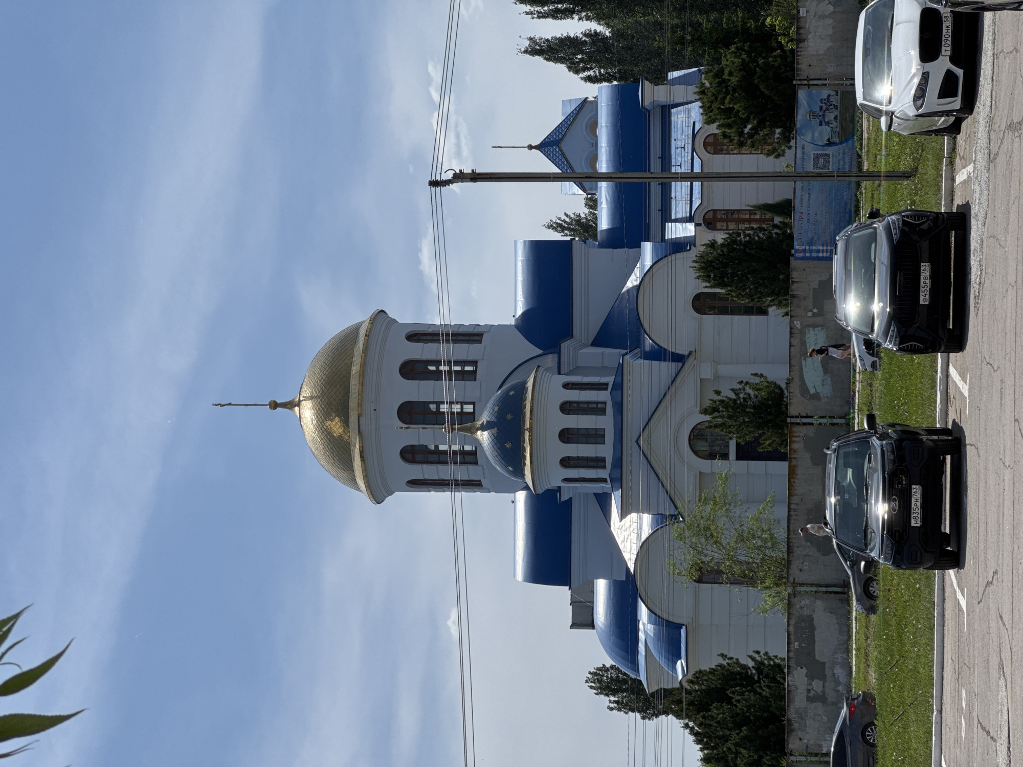 The image shows a white Orthodox church with multiple blue domes and a large gold dome, situated in what appears to be a city parking lot. Overhead power lines cross in front of the church. Several cars are parked in the lot, including a white car and a black SUV with a license plate visible as "H835PH 763". Vegetation and trees surround the church and parking lot. The sky is light blue with scattered clouds. The location is Unknown, Unknown.