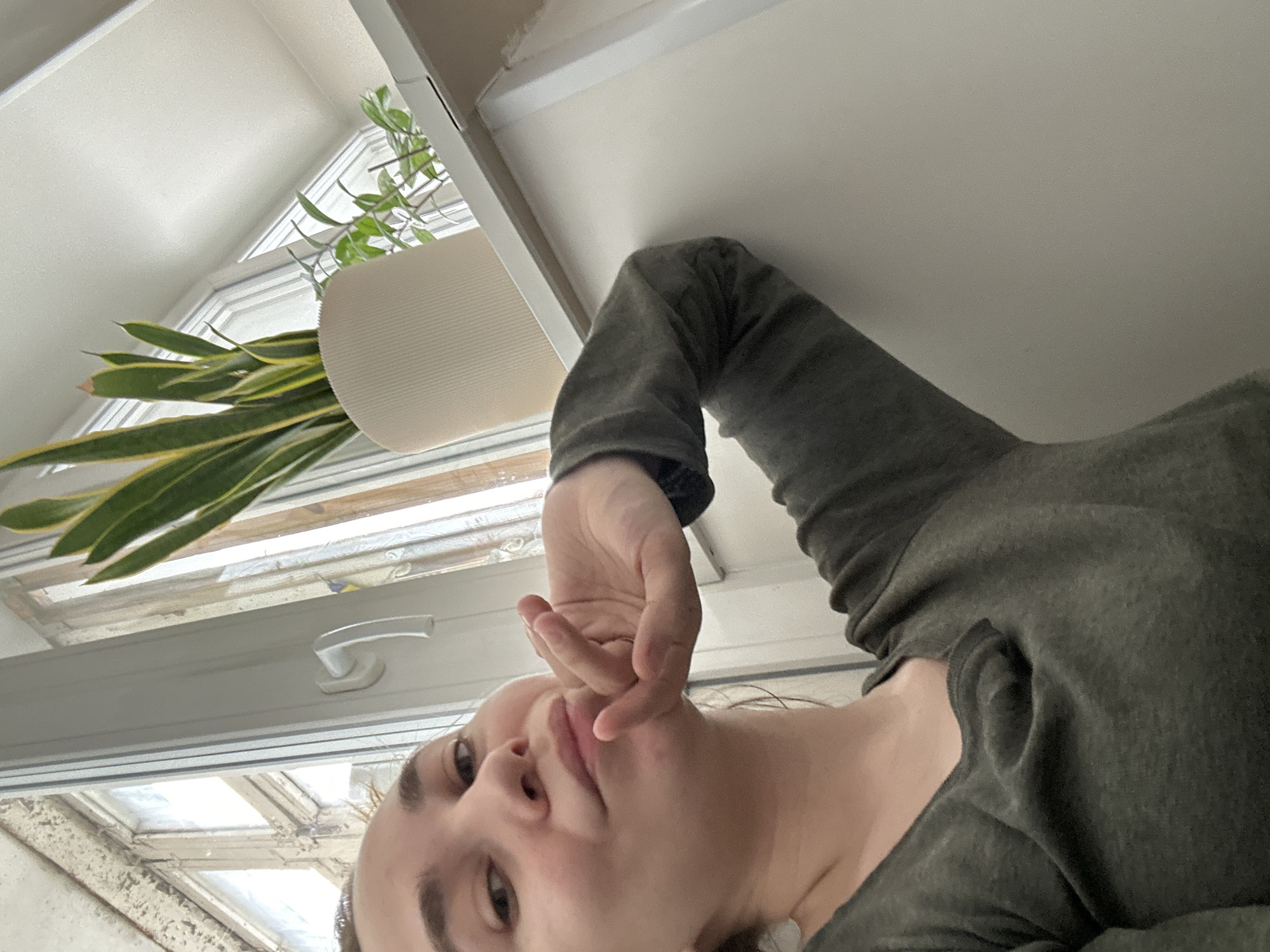 A low-angle, close-up shot features a person, likely female, with fair skin and dark hair, wearing an olive green long-sleeved shirt. Her face is partially visible, focusing on her eyes, nose, and mouth. Her right hand is raised near her face with fingers curled.
To the left of the person, a window with a white frame and handle is visible. Through the window, a plant in a white pot can be seen. The location seems to be indoors.
The city of Uva, Russia is not discernable from the image. There is no visual indicator of the city, its architecture, or its environment.