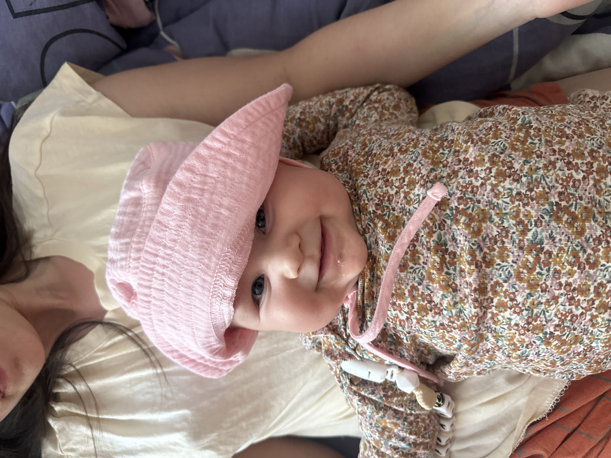 A baby wearing a pink hat and floral-patterned clothing is lying on a light-colored surface. A woman is partially visible to the left of the baby. The background includes bedding. There is no information in the image to suggest the location is Chaikovsky, Russia.