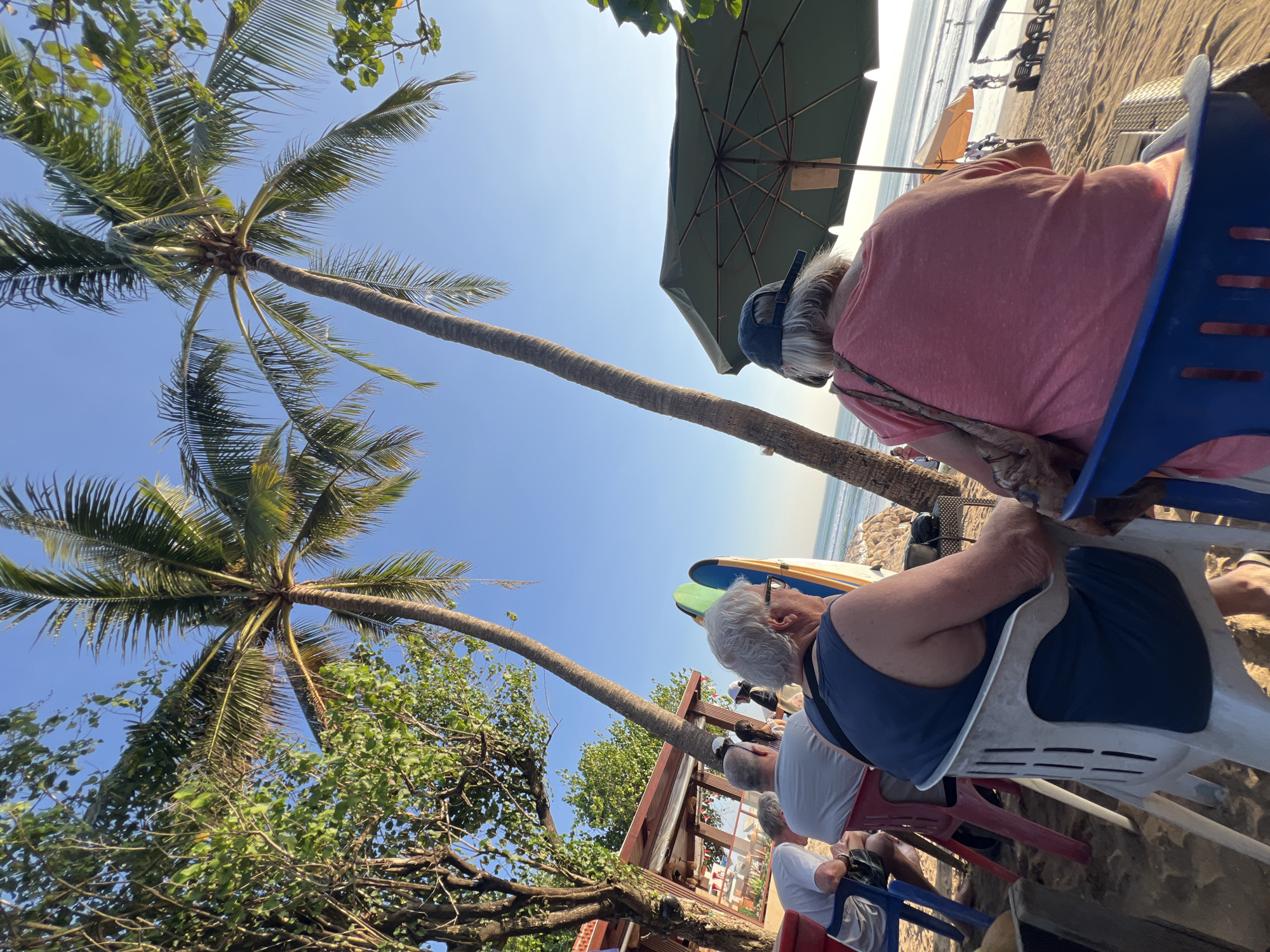 The image depicts a beach scene in Kuta, Indonesia, characterized by palm trees and clear blue skies. In the foreground, several people are seated on chairs on the sandy beach, facing the ocean. Two elderly individuals are prominently featured, viewed from behind. The woman on the right wears a pink t-shirt and a blue baseball cap, with a patterned bag slung over her shoulder. The man next to her, on the left, is wearing a dark blue tank top and glasses. In the background, the ocean is visible with small waves and distant figures on the beach and in the water. Surfboards are leaning against a structure behind the seated individuals. The overall setting suggests a relaxed atmosphere at a beachside establishment.