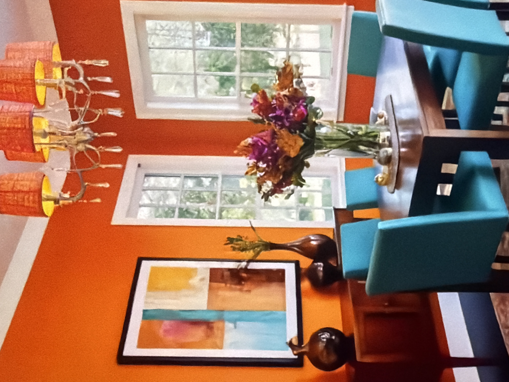 An interior dining room scene is captured. The walls are painted a vibrant orange. A dark wood dining table is set with teal chairs. On the table, a glass vase holds a colorful floral arrangement. To the left of the table, a dark wood cabinet displays two decorative vases and a framed abstract painting. Two windows, divided into panes, are visible on the back wall, revealing greenery outside. A multi-tiered chandelier with orange lampshades hangs from the ceiling. The floor is a dark color, possibly wood or tile.