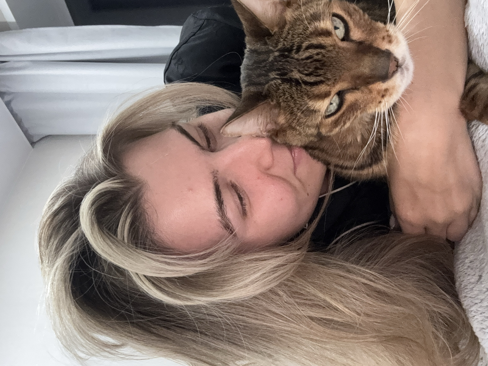 A close-up, slightly angled photograph shows a woman and a Bengal cat in an intimate pose. The woman, fair-skinned with closed eyes and long, blonde hair streaked with darker roots, is lying down. Her face is partially obscured by her hair and the cat's head. The Bengal cat, with its distinctive spotted brown and black coat, is positioned on top of the woman, its head resting near her face. Its eyes, green, are visible. The cat is seemingly interacting with the woman, possibly nuzzling or licking her face. The background includes a hint of a dark-colored garment, possibly clothing, and a gray textured fabric, potentially a blanket or pillow. The location is unknown.