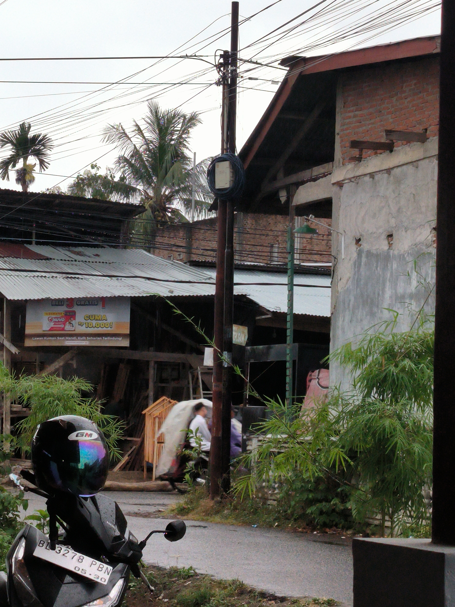 This image depicts an overcast street scene in Lhokseumawe, Indonesia, capturing a slice of urban daily life. In the foreground, a black Honda scooter is parked, facing slightly left, with a black GM helmet featuring an iridescent visor resting on its handlebars. The scooter's license plate reads "BL 3278 PBN," with "05.30" indicating a registration expiry. Lush green bamboo plants and other foliage line the left side of the damp asphalt road, suggesting recent rainfall.
In the mid-ground, a tall utility pole heavily laden with numerous electrical wires dominates the center, stretching towards an overcast sky. To the pole's left, a building with a corrugated metal roof displays a large advertising banner for "NUVO," stating "BELI GRATIS" (Buy Get Free), "CUMA Rp 10.000!" (Only Rp 10,000!), and "Lawan Kuman Saat Mandi, Kulit Seharian Terlindungi" (Fight Germs When Bathing, Skin Protected All Day Long). Two blurry figures are visible further back, one appearing to be on a scooter or walking, near a small, light-colored wooden structure.
To the right, a building constructed of red brick and partially covered with grey plaster shows an exposed wooden roof frame. A green street light fixture extends from a pole near this building. The background features several palm trees and an intricate web of power lines crisscrossing the bright, hazy sky. The scene conveys a typical daytime atmosphere in a vibrant Southeast Asian setting.