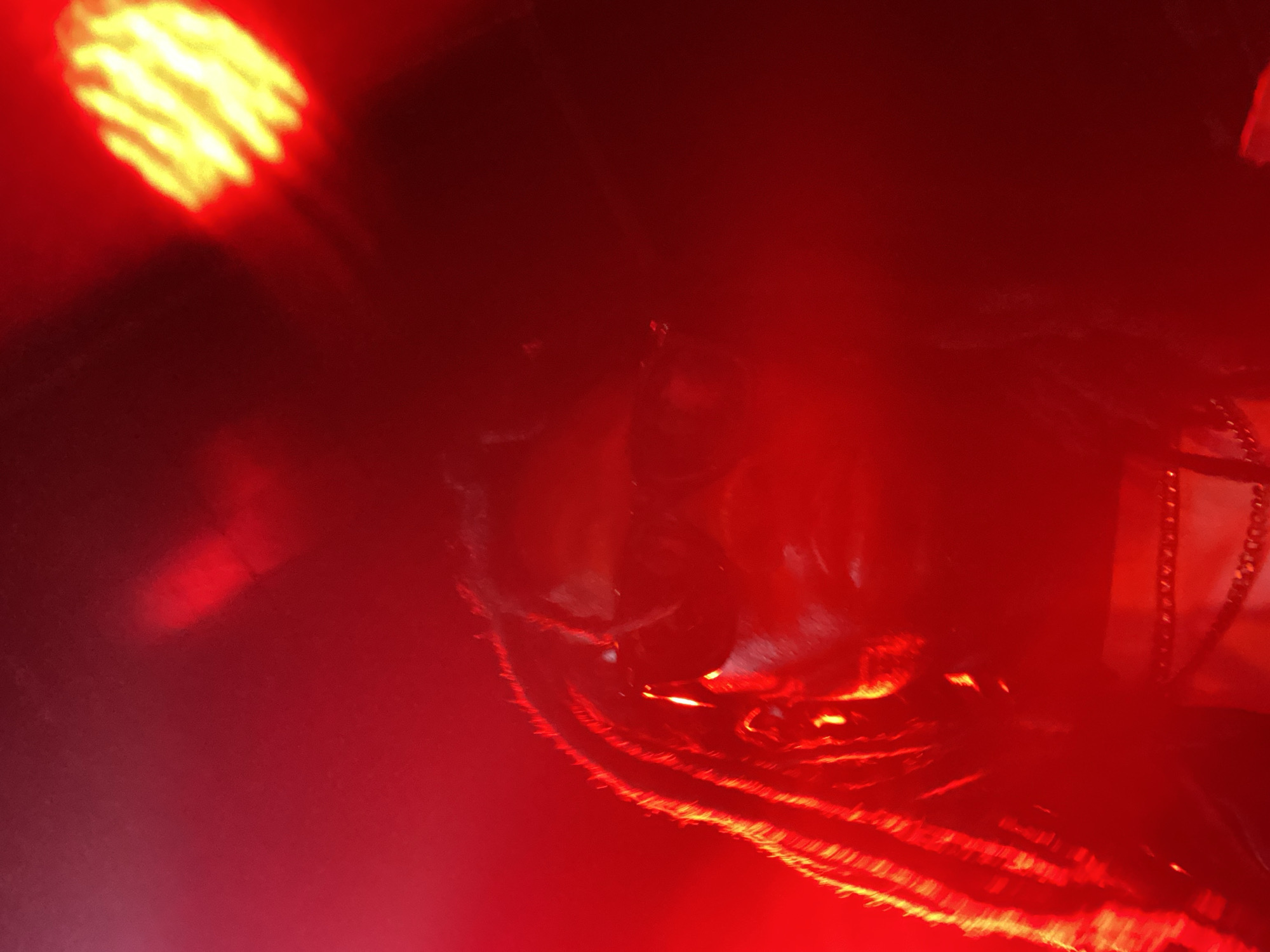 The image captures a dimly lit, red-hued environment, likely a performance venue or nightclub. In the foreground, a person with dreadlocks is partially visible, illuminated by bright stage lights. The person appears to be wearing dark clothing, possibly with some metallic or reflective accents around the neck. The background is mostly obscured by the red lighting and shadows, but a bright yellow spotlight with a textured pattern is visible in the upper right quadrant, suggesting the presence of stage lighting equipment. The overall atmosphere is dark, intense, and suggestive of a live event. There is no visual information in the image that can confirm a specific location in Abuja, Nigeria.