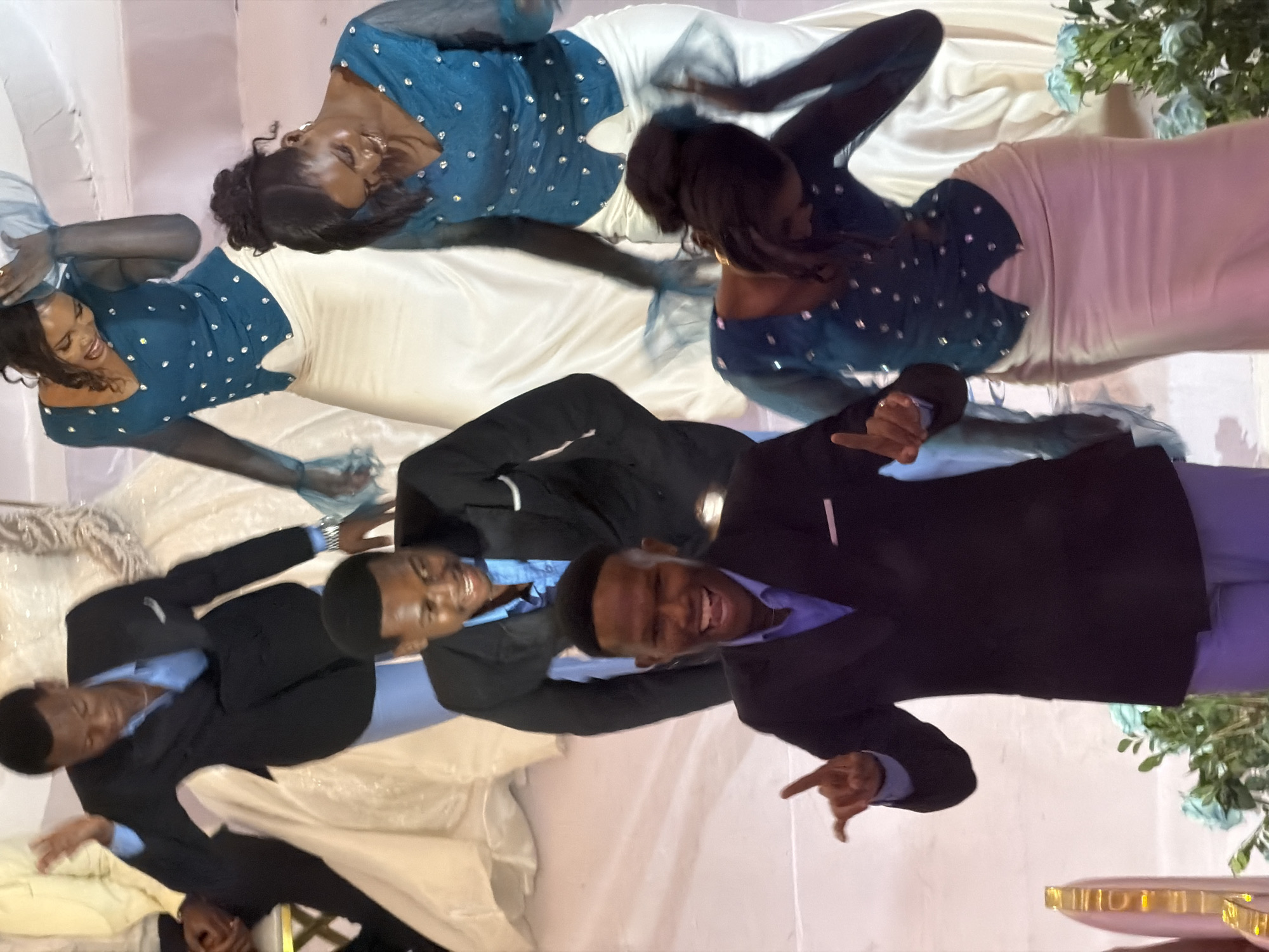 The image captures a moment of celebration, likely a wedding or formal event, taking place indoors with a light-colored backdrop. Five individuals are visible, dressed in formal attire. In the foreground, a man in a dark suit and light blue shirt is energetically dancing, with his mouth open in a wide smile and his right hand raised, fingers pointed upwards. Behind him and slightly to his left, another man in a similar suit and blue shirt is also smiling and looking towards the camera, with his arms extended.
Further back, three women are engaged in the celebration. Two of them, positioned to the right, are wearing dark teal tops adorned with rhinestones and long, flowing light-colored skirts. One of these women has her head tilted down, her hair pulled back, and appears to be dancing. The other woman next to her is also dancing, with her head turned to the side and her arms in motion. A third woman, on the left, is partially visible and appears to be wearing a white wedding gown, suggesting a wedding context. The environment seems to be decorated with light-colored fabric drapes and some light blue floral arrangements. The overall atmosphere is joyful and celebratory. The image was taken in Dar es Salaam, Tanzania.