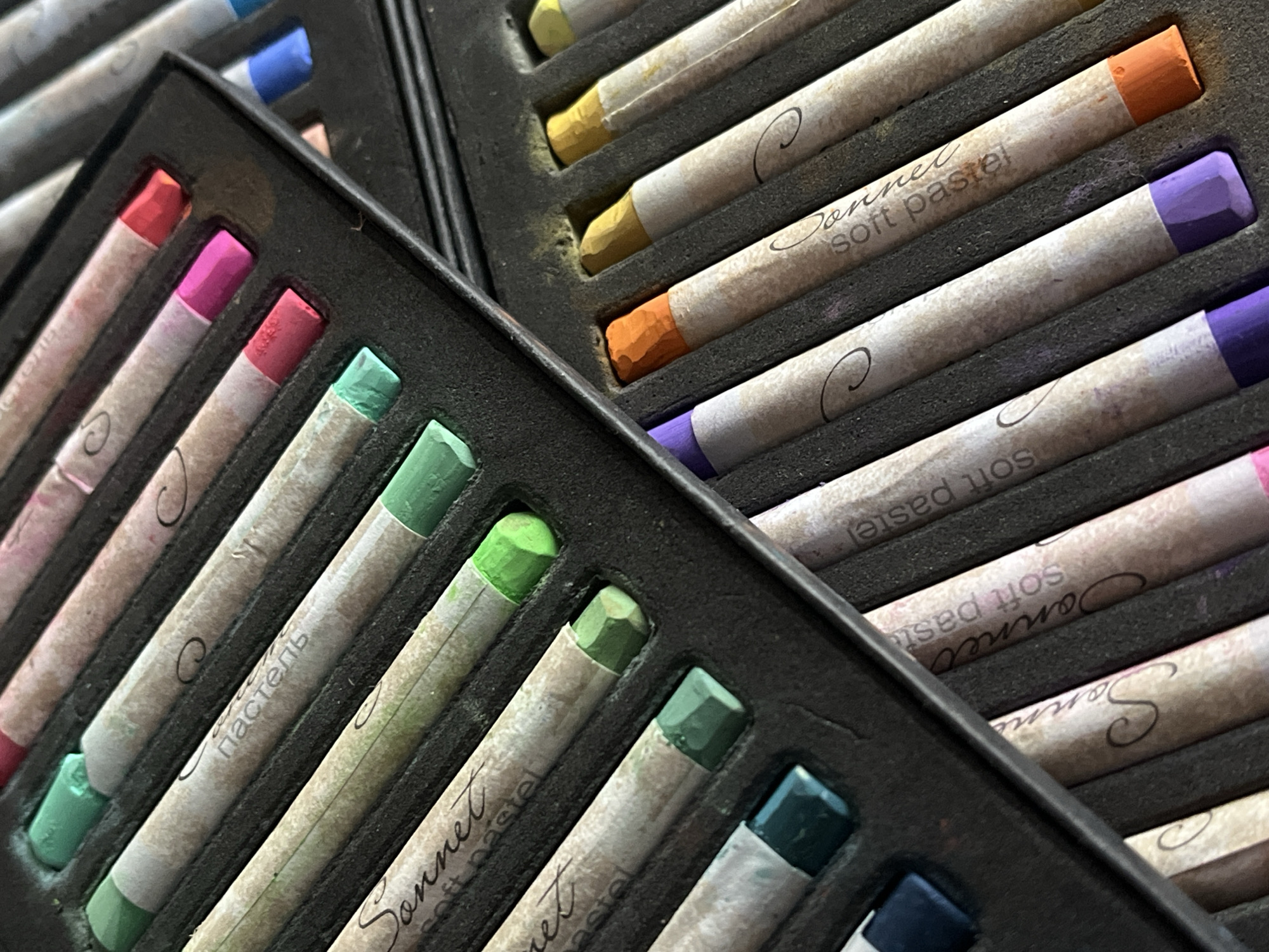 The image shows a close-up of two partially visible boxes of "Sonnet soft pastel" crayons. The box in the foreground contains crayons in various shades of green, pink, and red. The box in the background contains crayons in shades of yellow, orange, and purple. The brand name "Sonnet soft pastel" is printed on the crayons. The setting appears to be indoors, but the location in the city of Unknown, Unknown cannot be determined from the image. There are no people or actions visible.